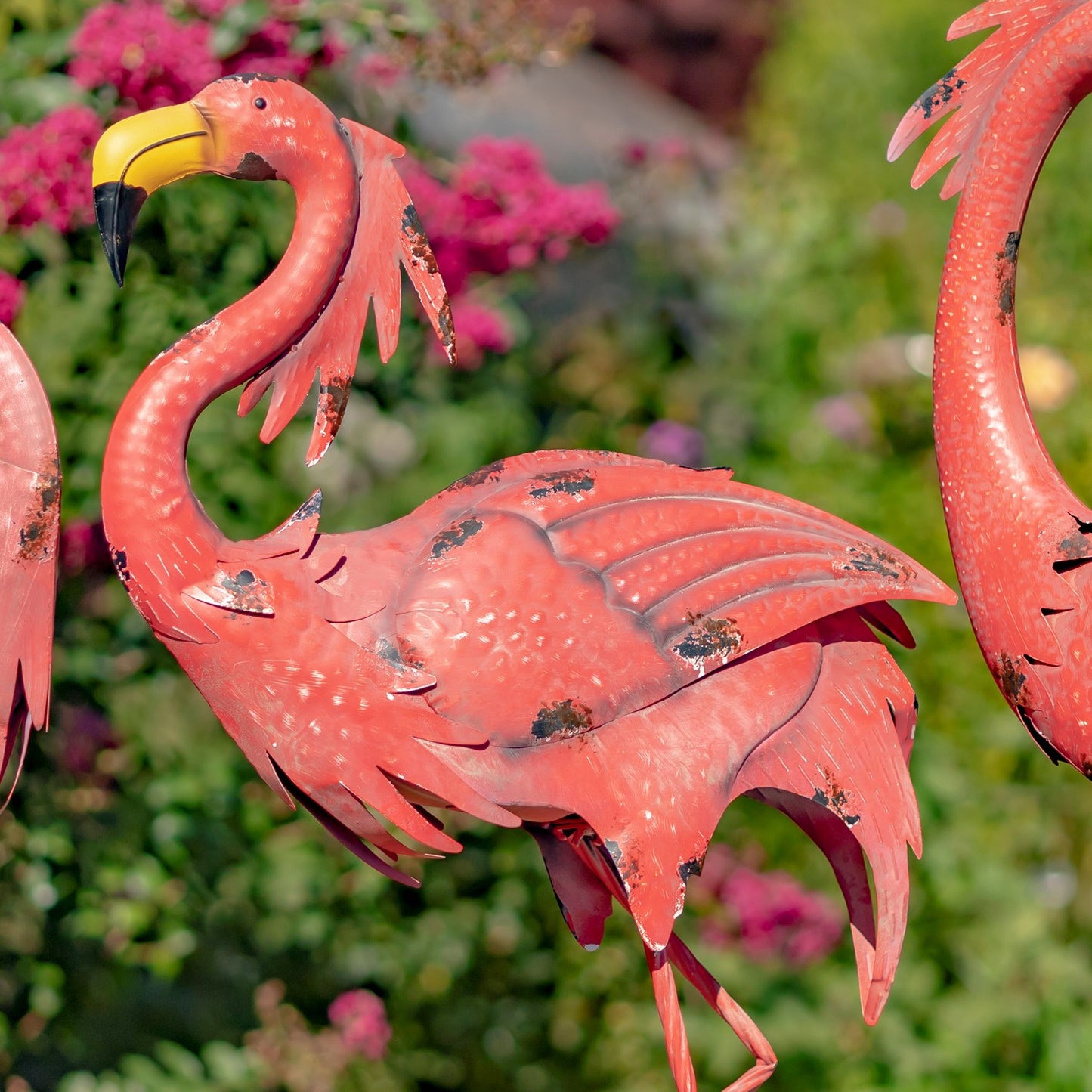 A decorative metal flamingo garden statue in a bright pinkish-red finish, shown outdoors with a blurred green garden and pink flowers in the background.