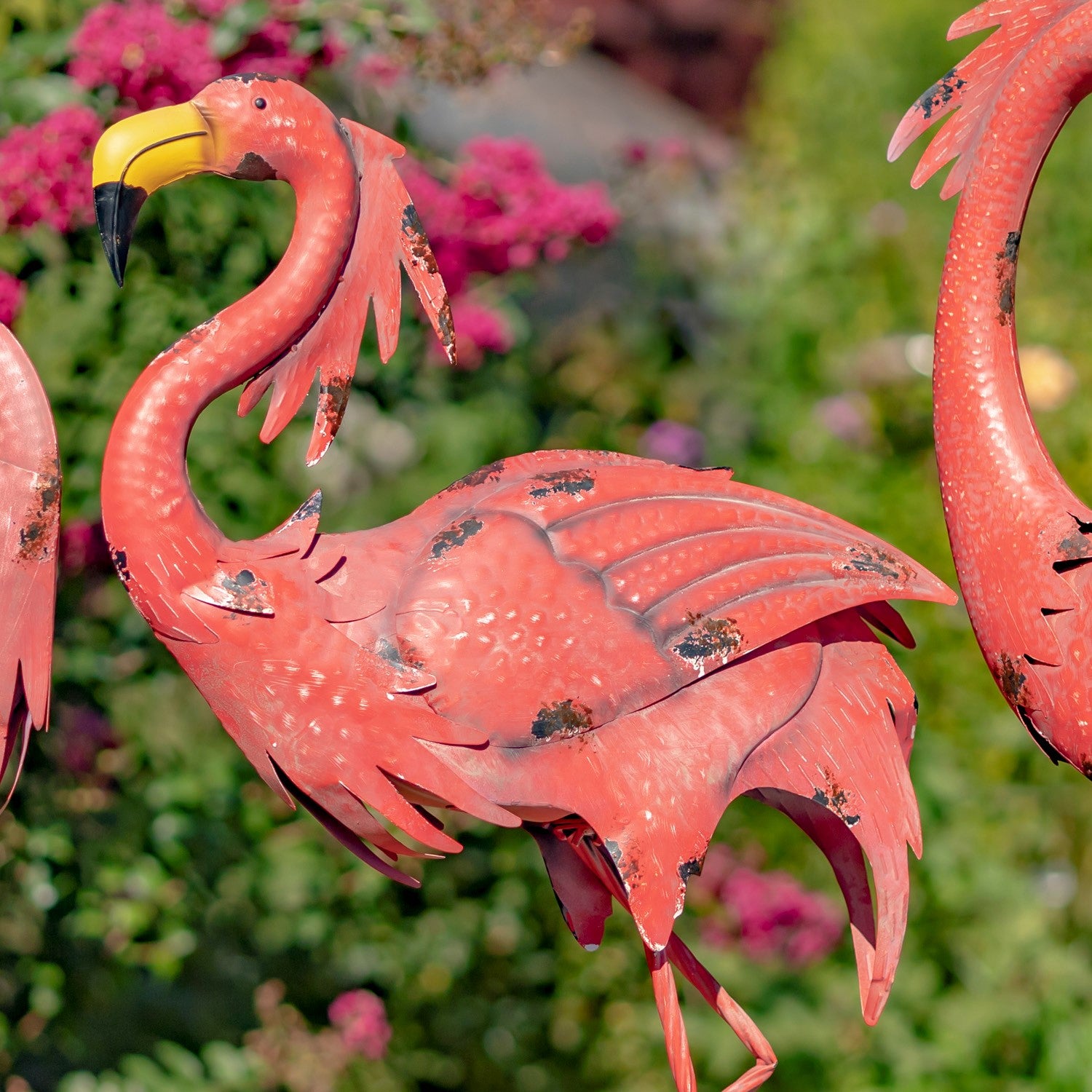A decorative metal flamingo garden statue in a bright pinkish-red finish, shown outdoors with a blurred green garden and pink flowers in the background.