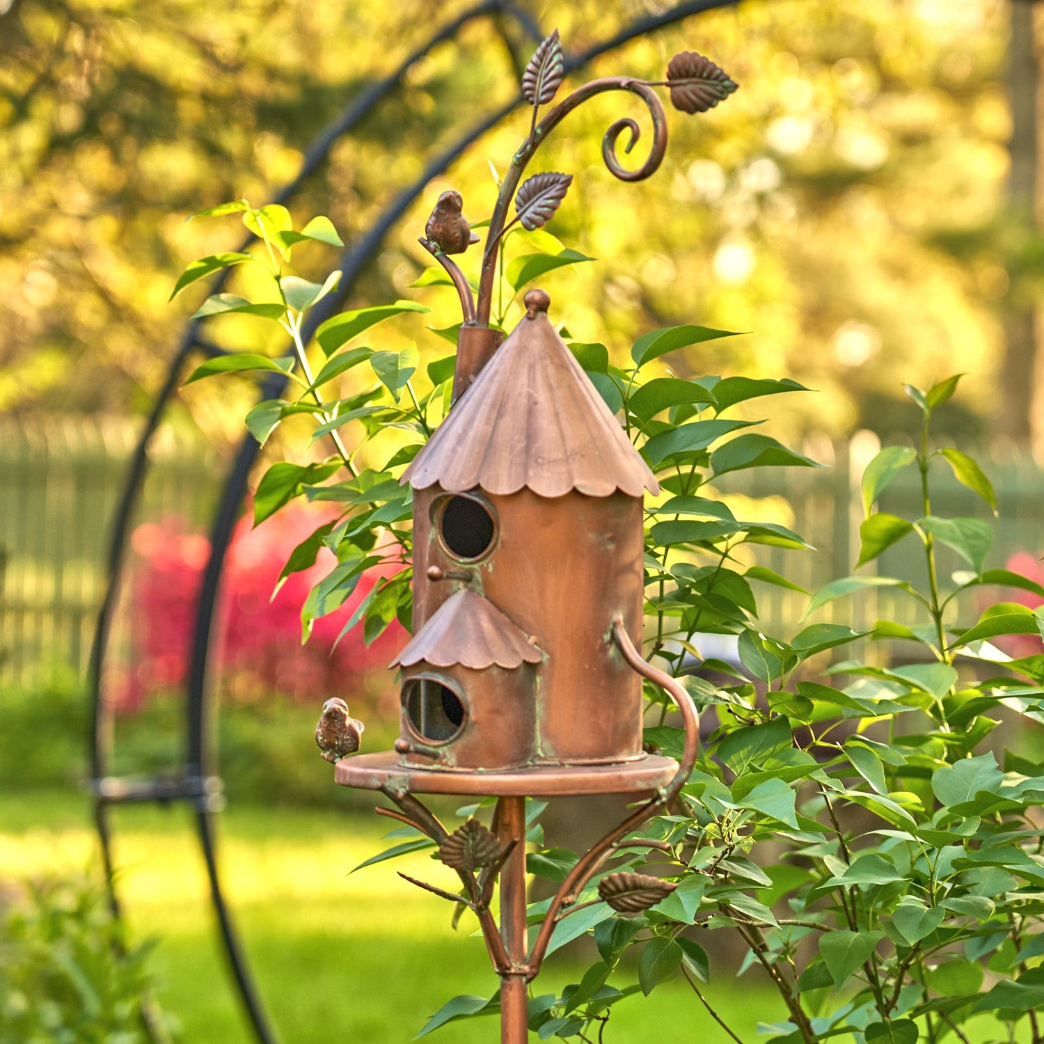 A decorative copper birdhouse with a small bird perched on it, mounted on a matching metal garden stand in a sunlit garden with green foliage and blurred background trees.