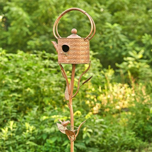 A decorative metal garden stake shaped like a birdhouse with a watering can shape, featuring a small bird perched at the bottom, set against a blurred green garden background.