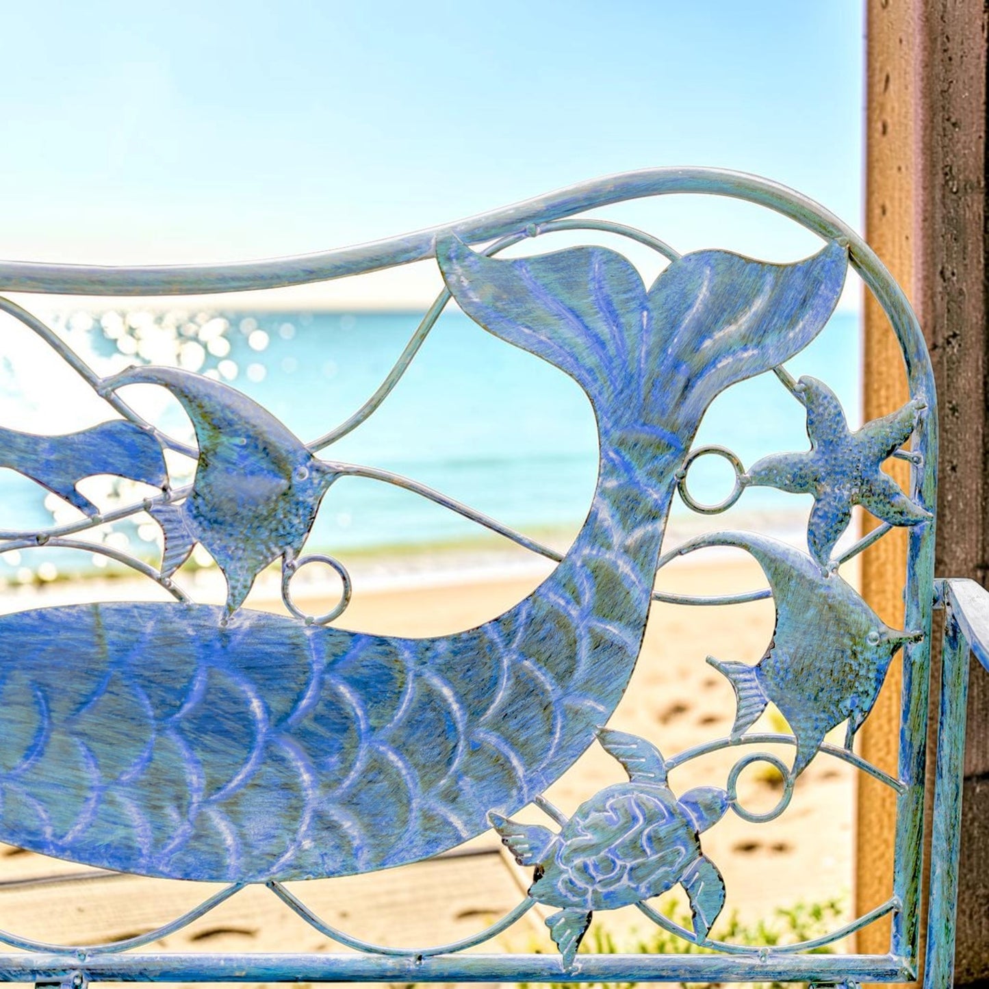 A decorative metal garden gate featuring a mermaid and sea life design, including a mermaid tail, fish, starfish, and turtle, painted in a distressed light blue and white finish. The gate is shown outdoors with a blurred beach and ocean background.