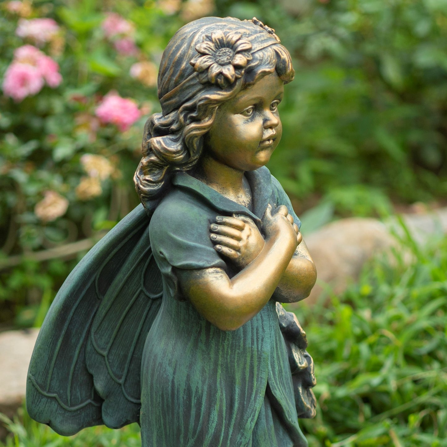 Bronze-toned garden statue of a young winged child with a flower in the hair, standing in a garden with green foliage and pink flowers in the background.