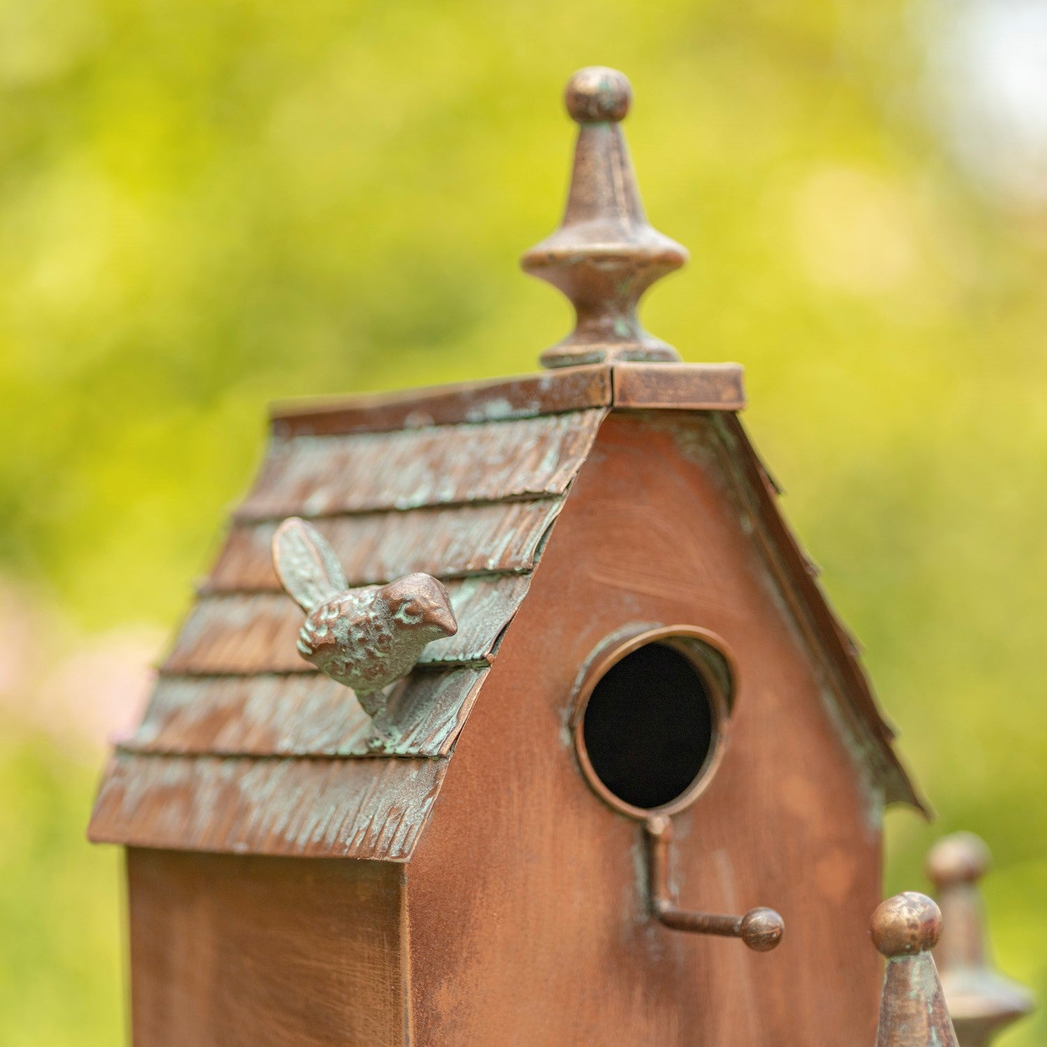 A multi-tiered birdhouse with a rustic brown and bronze finish, mounted on a decorative metal stand with leaf details, set against a blurred green garden background with pink flowers.