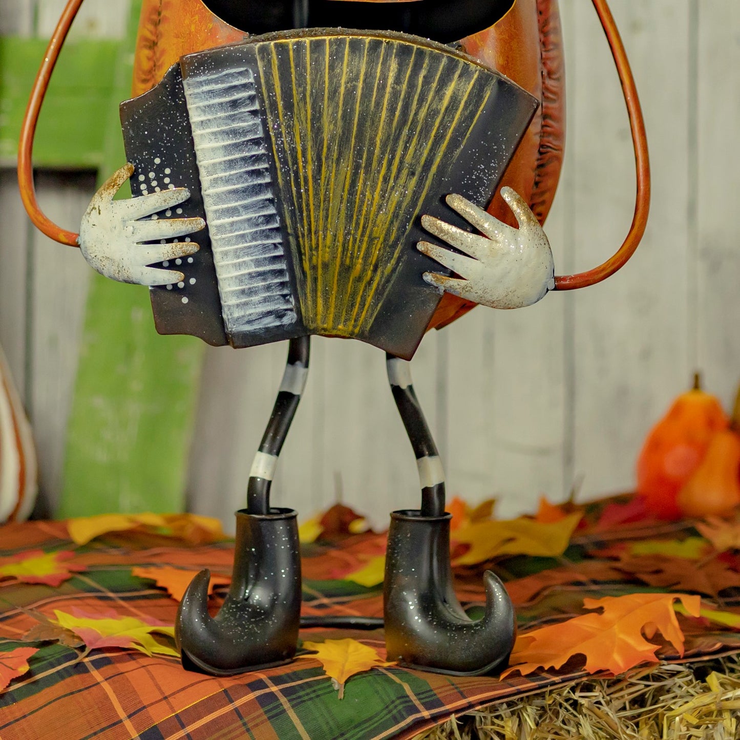A decorative metal pumpkin figurine playing an accordion, standing on a plaid blanket with autumn leaves and small pumpkins in the background.