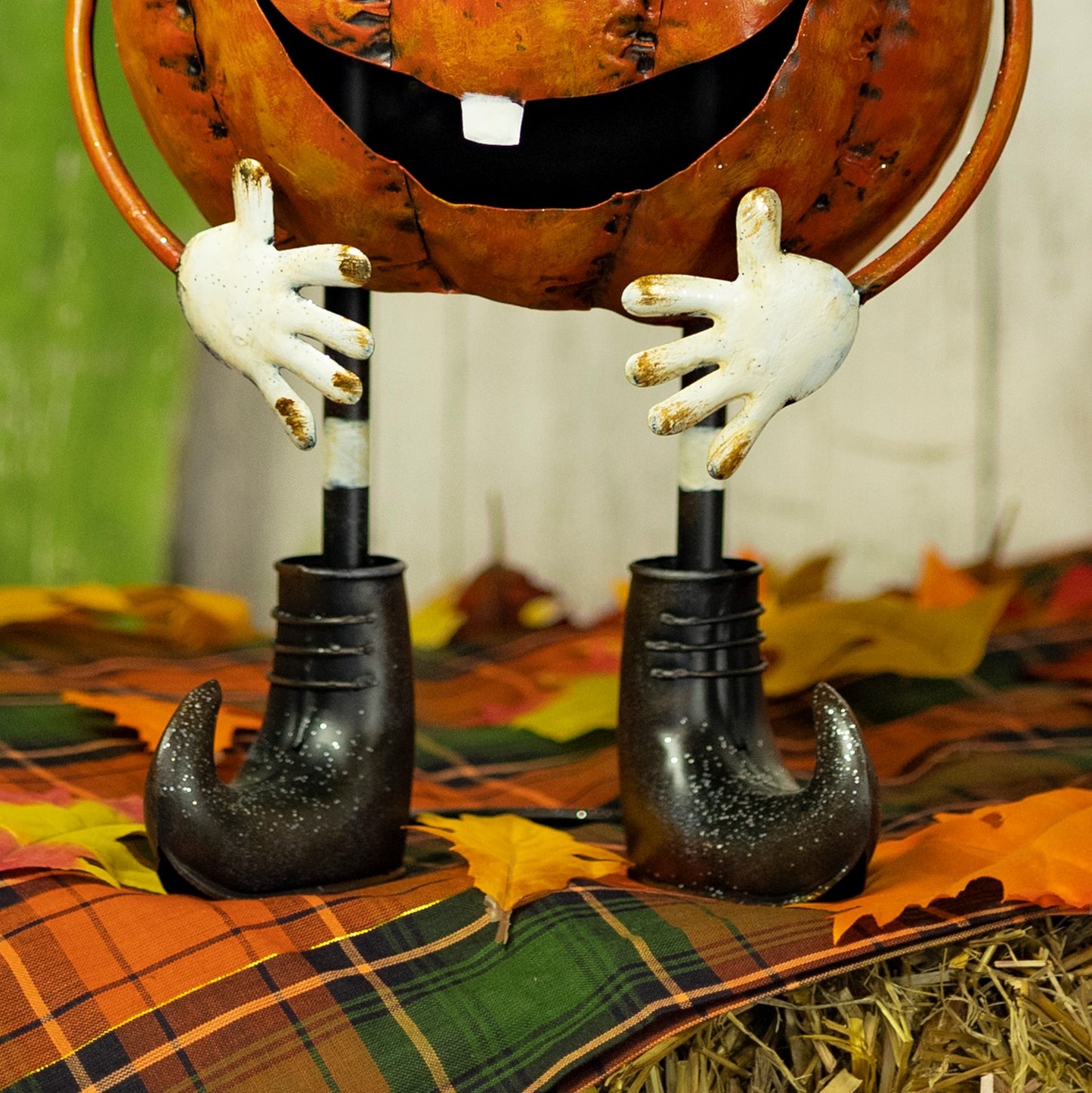 A Halloween decorative pumpkin figure with a large orange jack-o'-lantern head, white hands, and black boot-shaped legs, posed on a plaid blanket with autumn leaves.