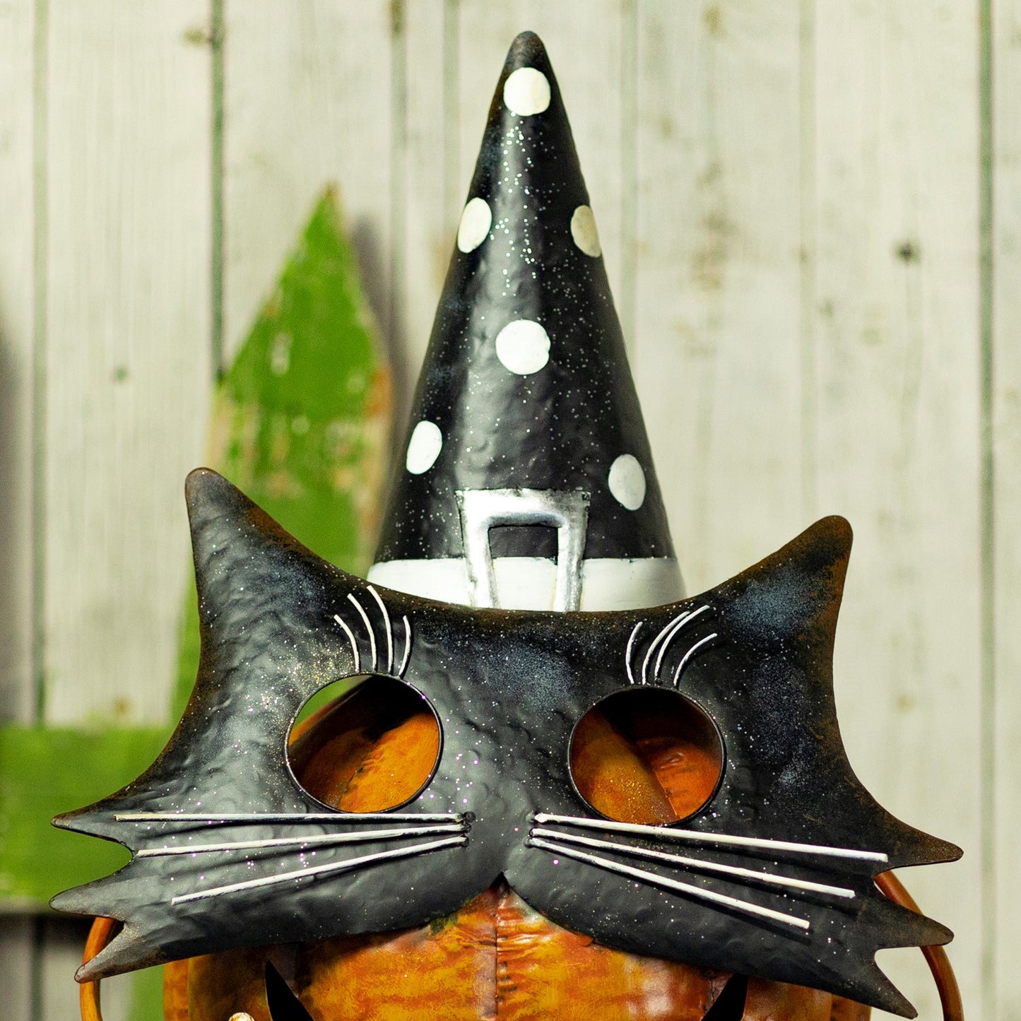 A black witch hat and black cat mask set with silver glitter accents, worn by a person holding a glowing orange pumpkin against a rustic white wood plank background.