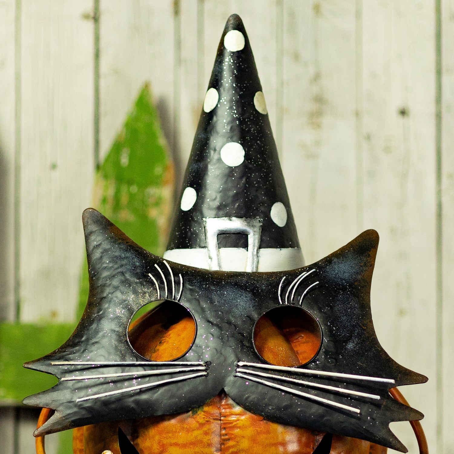 A black witch hat and black cat mask set with silver glitter accents, worn by a person holding a glowing orange pumpkin against a rustic white wood plank background.