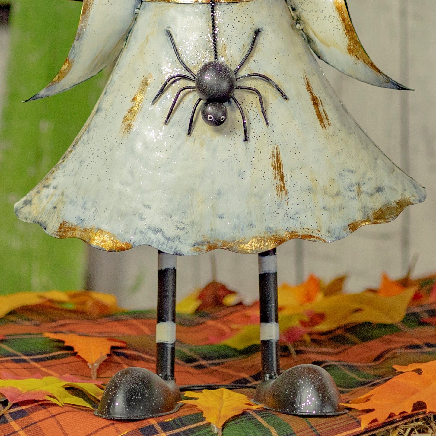 A decorative metal angel figurine with a distressed off-white dress, gold trim, and a hanging black spider ornament, standing on striped legs over a plaid autumn leaf surface.