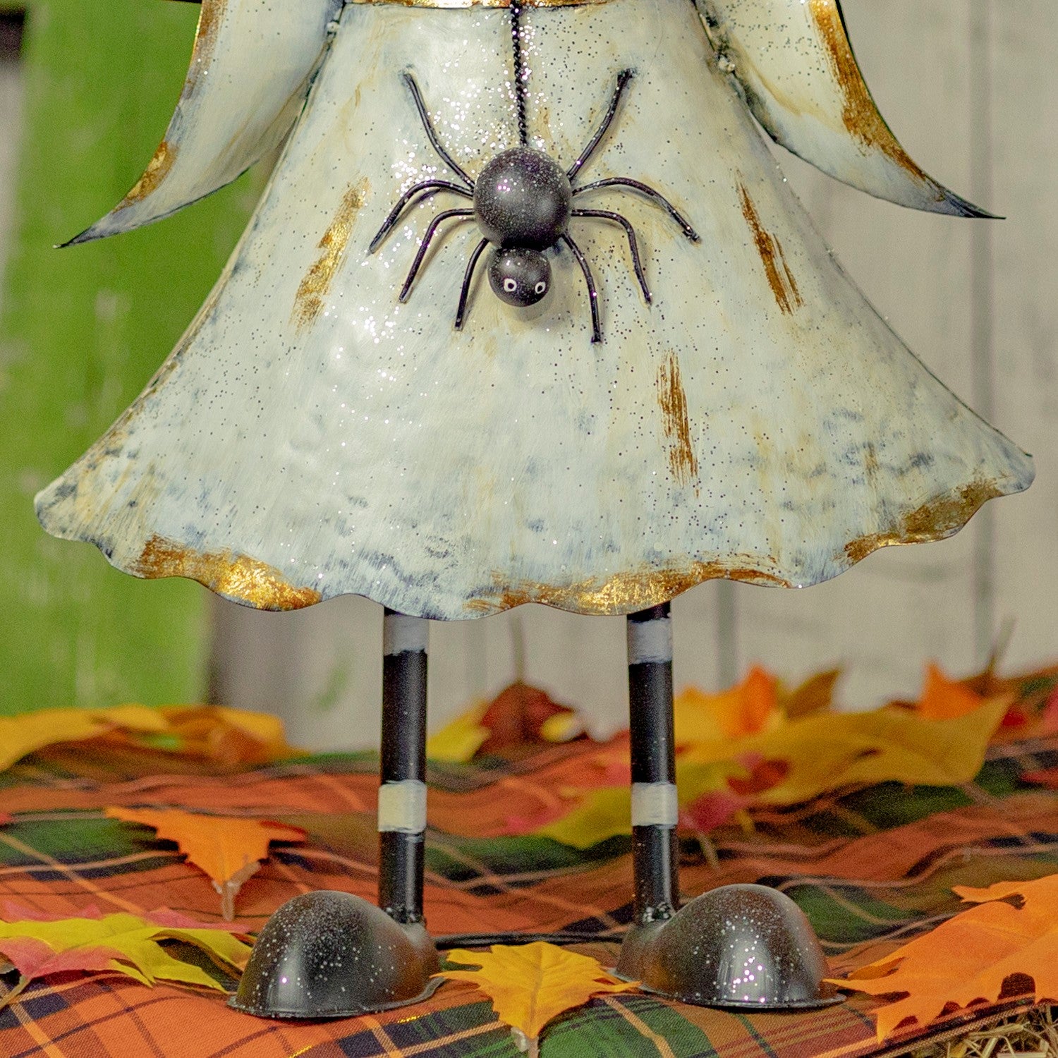 A decorative metal angel figurine with a distressed off-white dress, gold trim, and a hanging black spider ornament, standing on striped legs over a plaid autumn leaf surface.