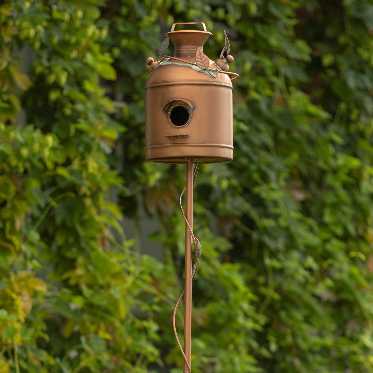 A rustic metal birdhouse designed like a vintage milk canister, mounted on a tall metal pole against a background of green ivy climbing a fence.