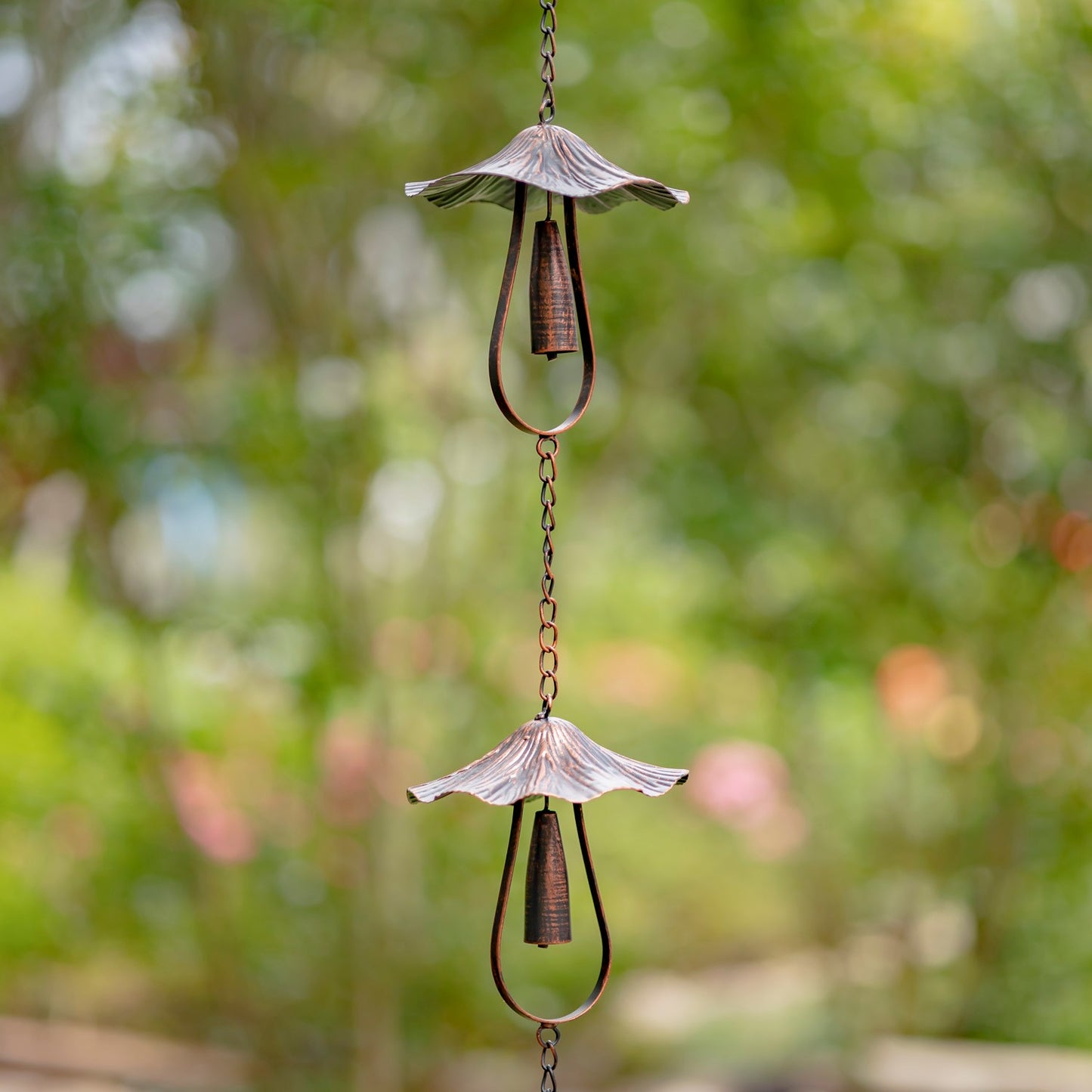 A hanging metal wind spinner with two decorative cup-like blades and dangling rings, photographed outdoors against a blurred green garden background.