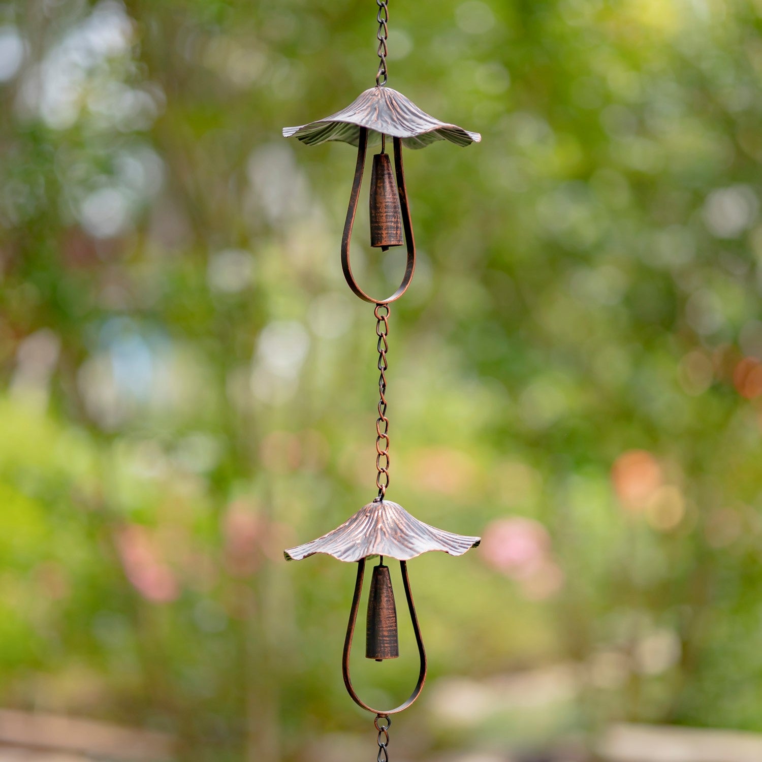A hanging metal wind spinner with two decorative cup-like blades and dangling rings, photographed outdoors against a blurred green garden background.
