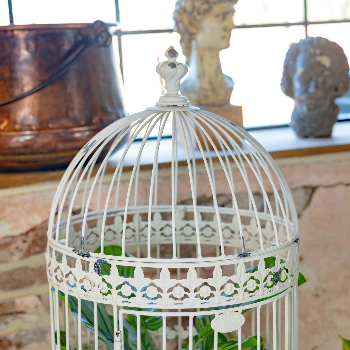 A white metal birdcage with decorative cutout detailing and a small finial on top, shown in a styled interior setting with blurred busts and a copper pot in the background.