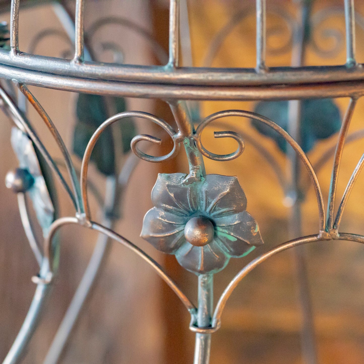 Close-up of an ornate metal stand or shelf with a distressed silver finish and a central flower-shaped decorative element, shown against a softly blurred warm brown background.