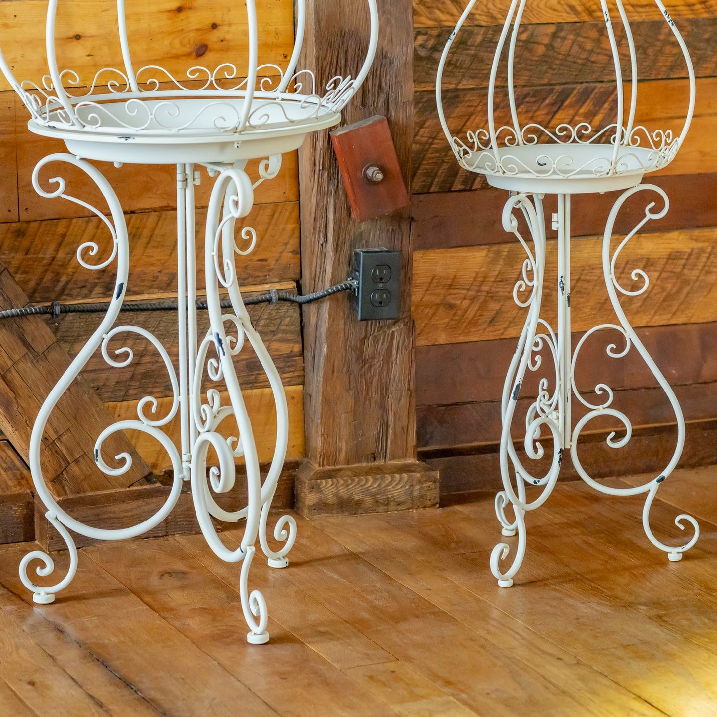 Two white wrought iron plant stands with ornate scrollwork legs, shown on a wooden floor against a rustic wood-paneled wall.