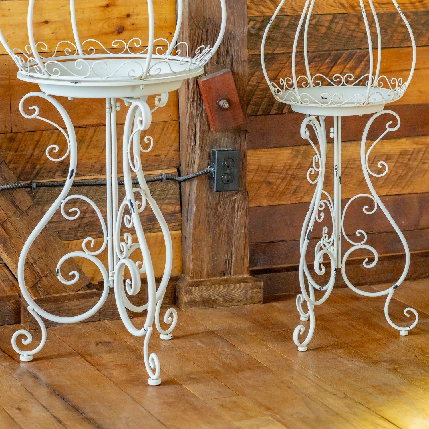Two white wrought iron plant stands with ornate scrollwork legs, shown on a wooden floor against a rustic wood-paneled wall.
