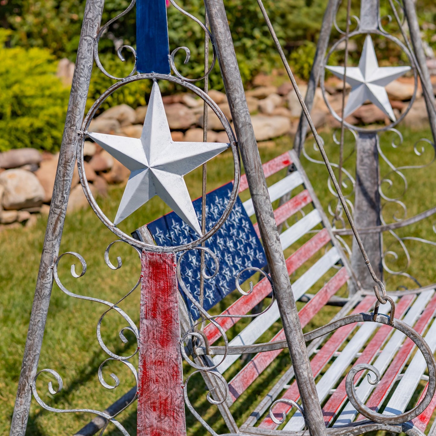 Patriotic metal swing bench with a distressed silver frame, white star decorations, and a red, white, and blue American flag design on a grassy lawn.