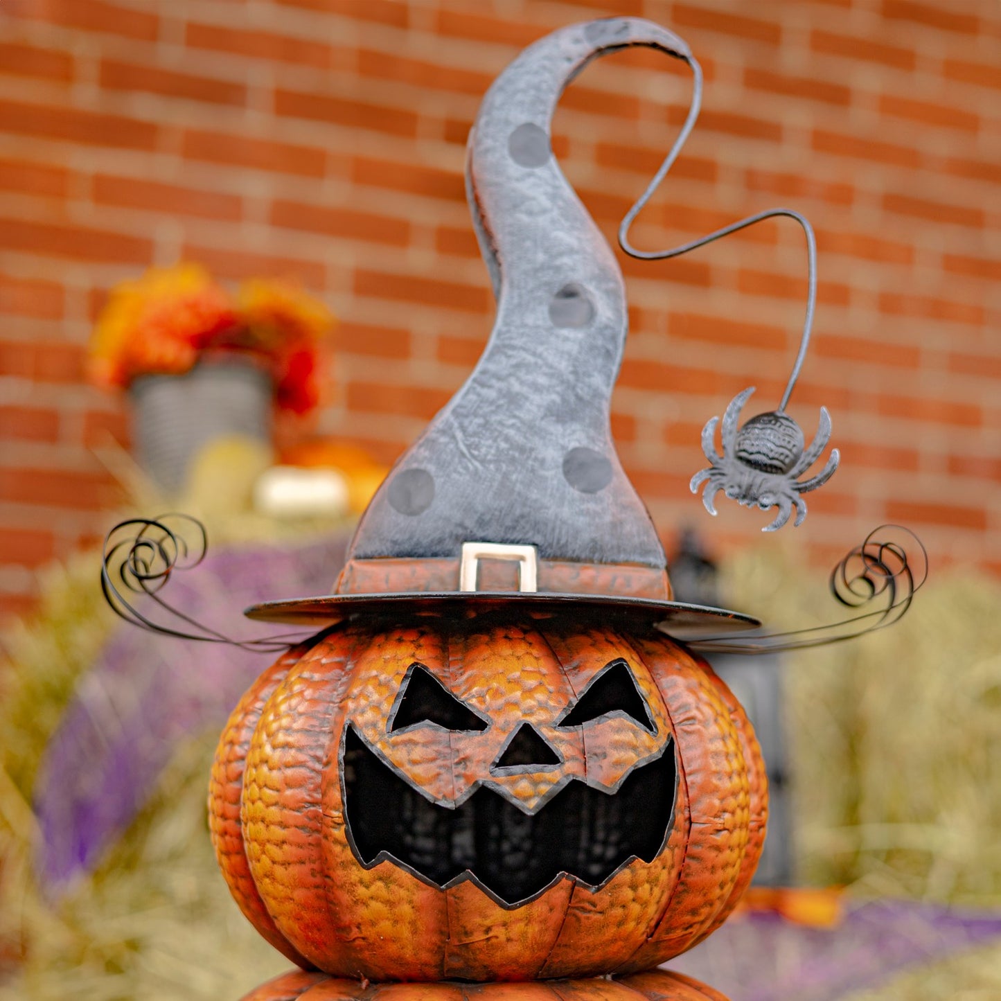 A decorative jack-o'-lantern pumpkin topped with a tall pointed witch hat and a small hanging spider ornament, set outdoors in front of a blurred brick wall.