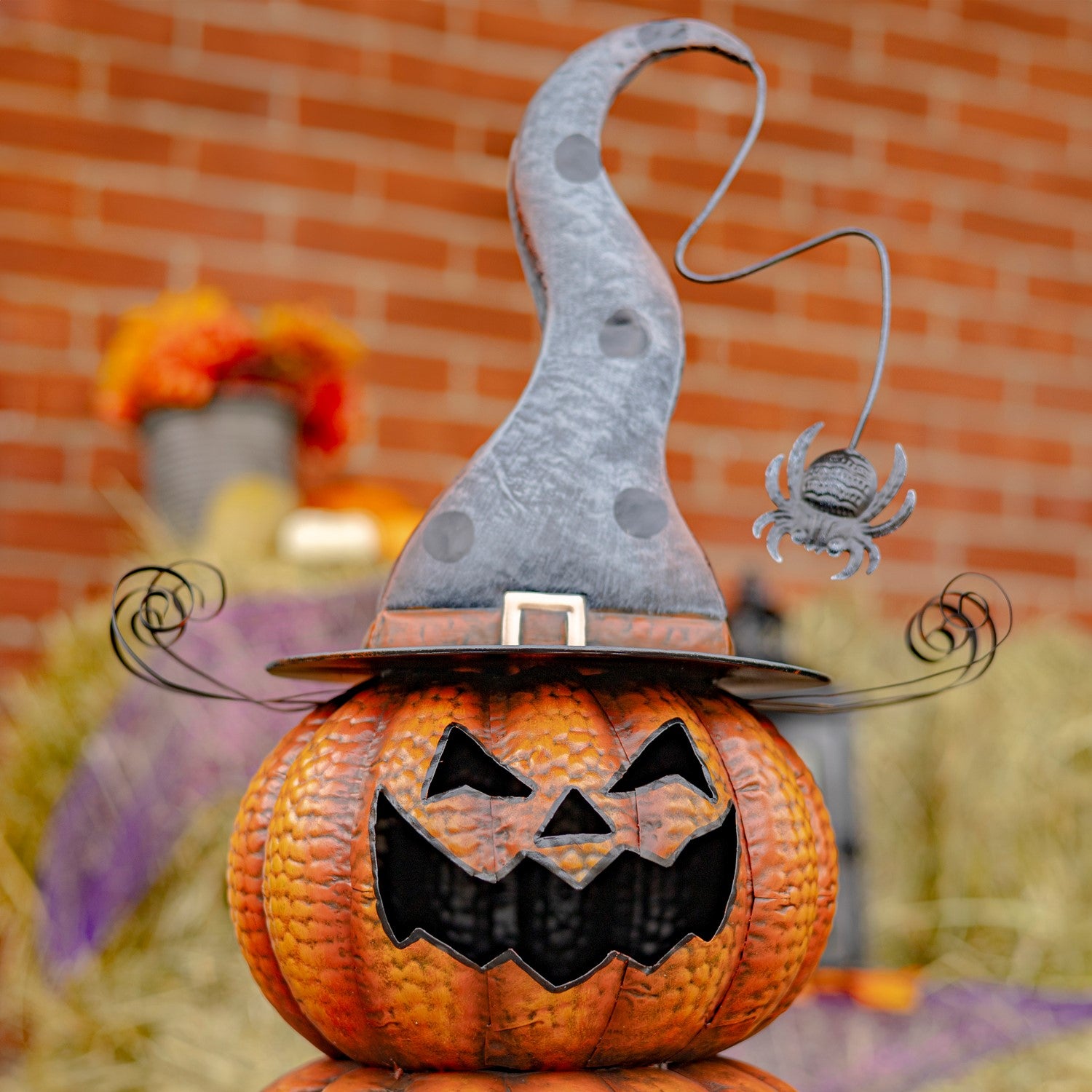 A decorative jack-o'-lantern pumpkin topped with a tall pointed witch hat and a small hanging spider ornament, set outdoors in front of a blurred brick wall.
