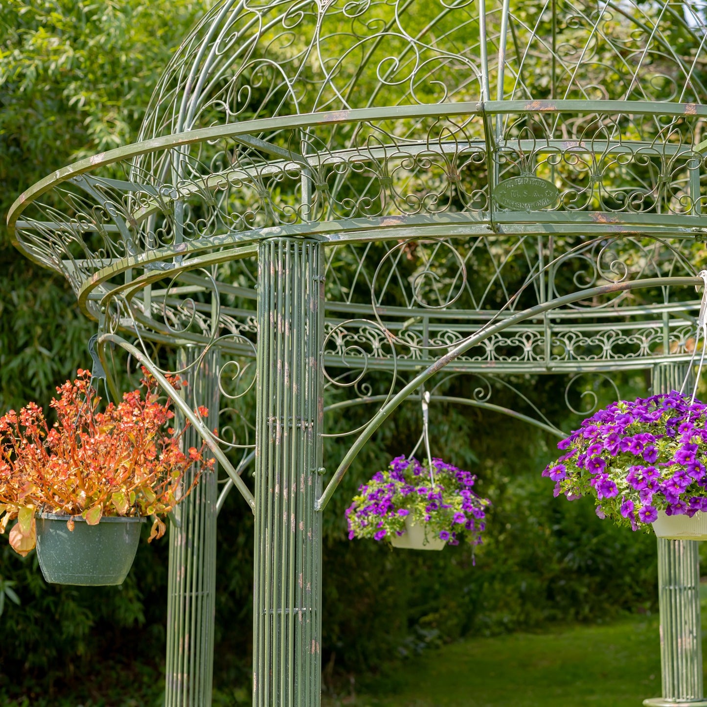A light sage green metal garden gazebo with decorative scrollwork, shown outdoors in a garden with three hanging flower baskets, including one with orange flowers and two with purple flowers.