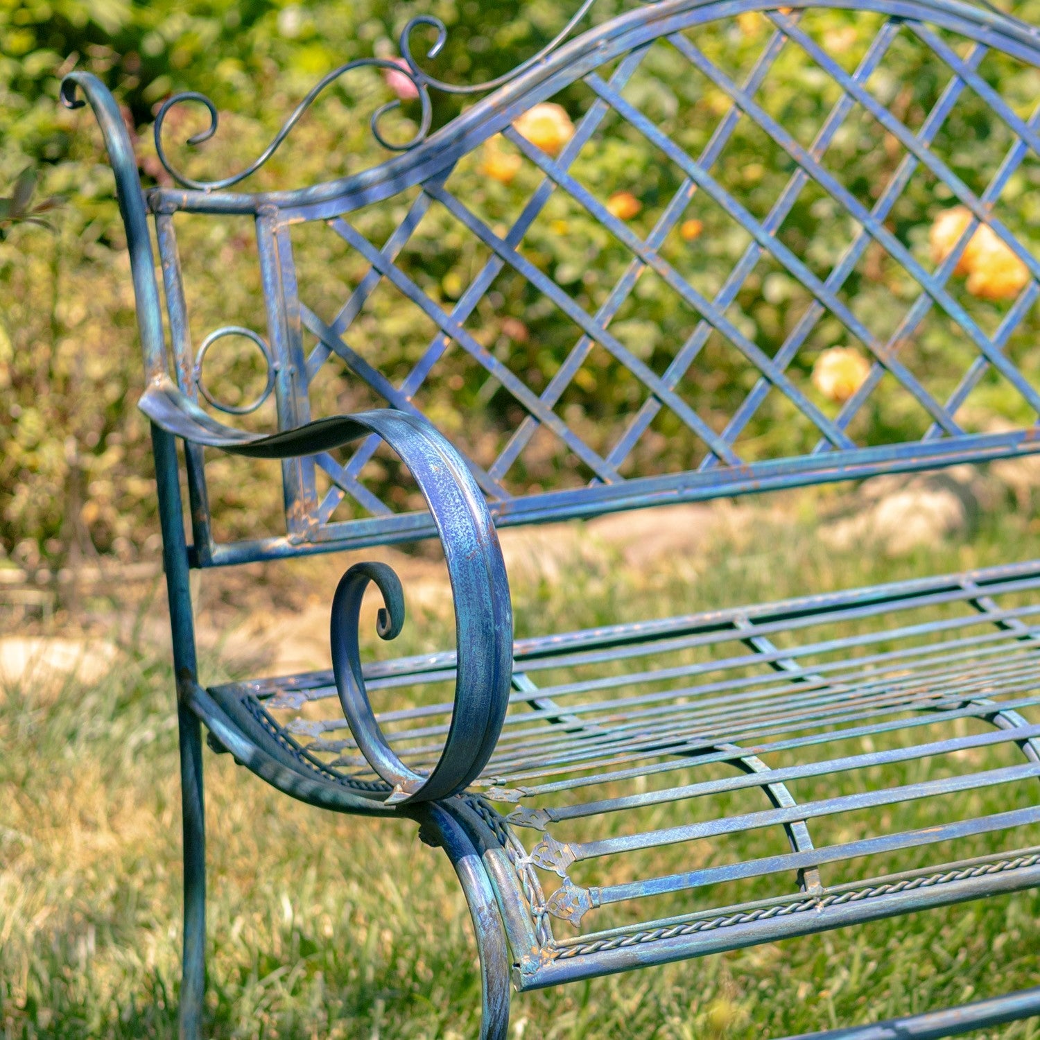 A close-up of an outdoor metal bench with a distressed blue-green finish, featuring scrollwork on the armrests and a lattice backrest, set in a grassy garden with blurred foliage.