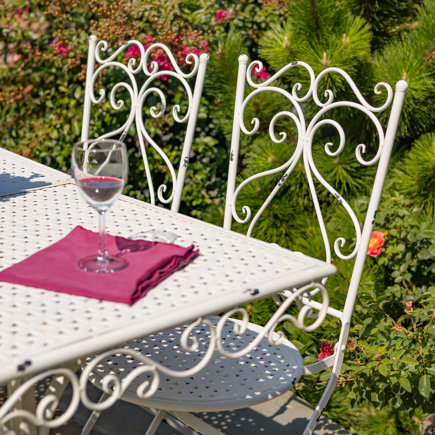 White metal garden table and chairs with a glass of red wine and pink napkin on a sunny day.