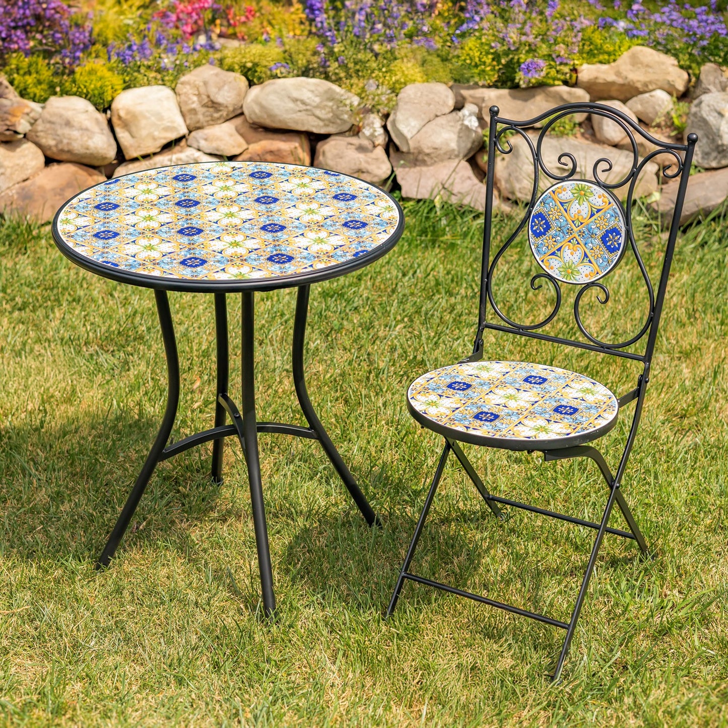 Outdoor bistro set with a round black metal table and matching folding chair featuring a colorful mosaic tile pattern on the tabletop and backrest, set on a green lawn with a stone garden bed in the background.