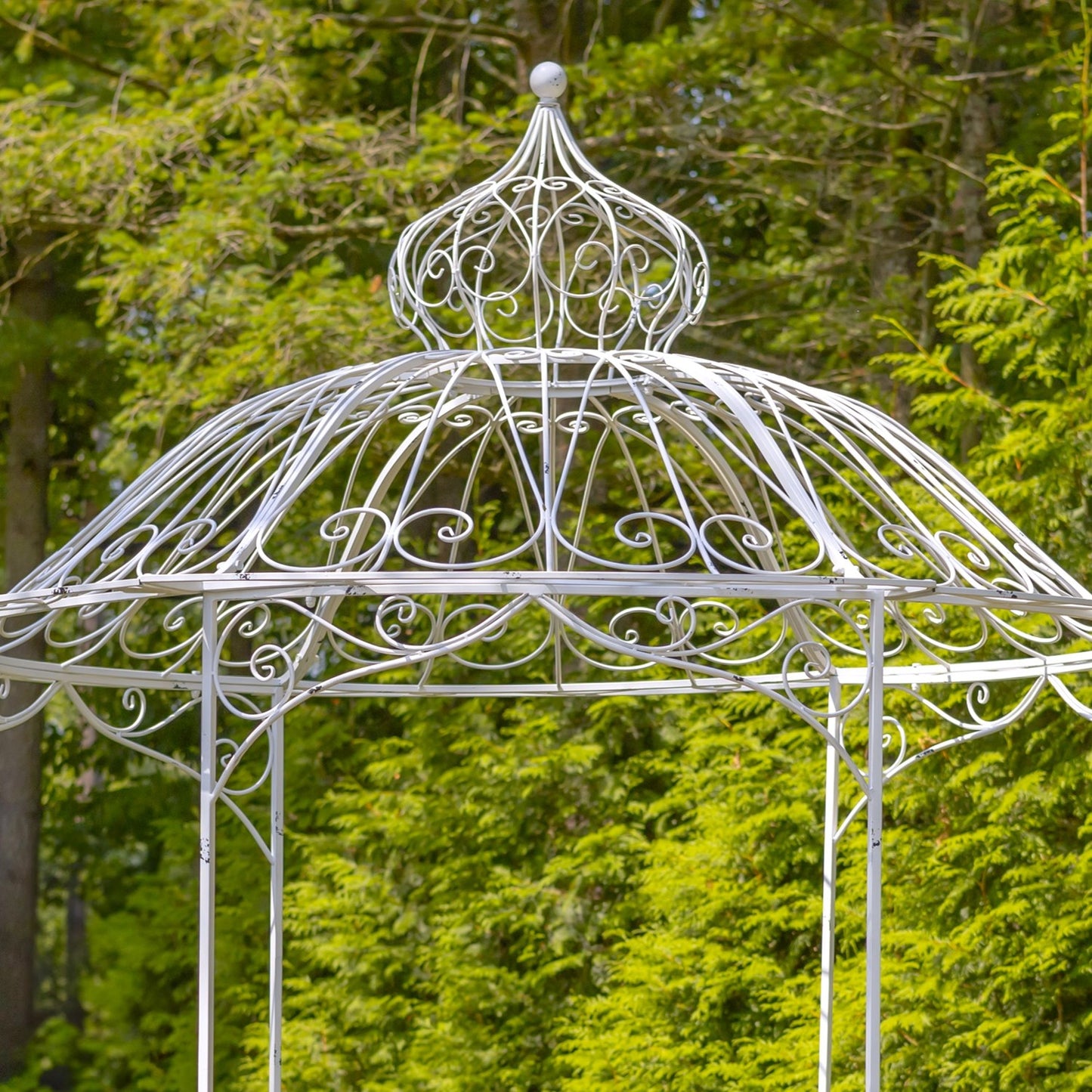 A white wrought iron garden gazebo with an ornate domed roof and decorative scrollwork, set against a background of lush green trees.