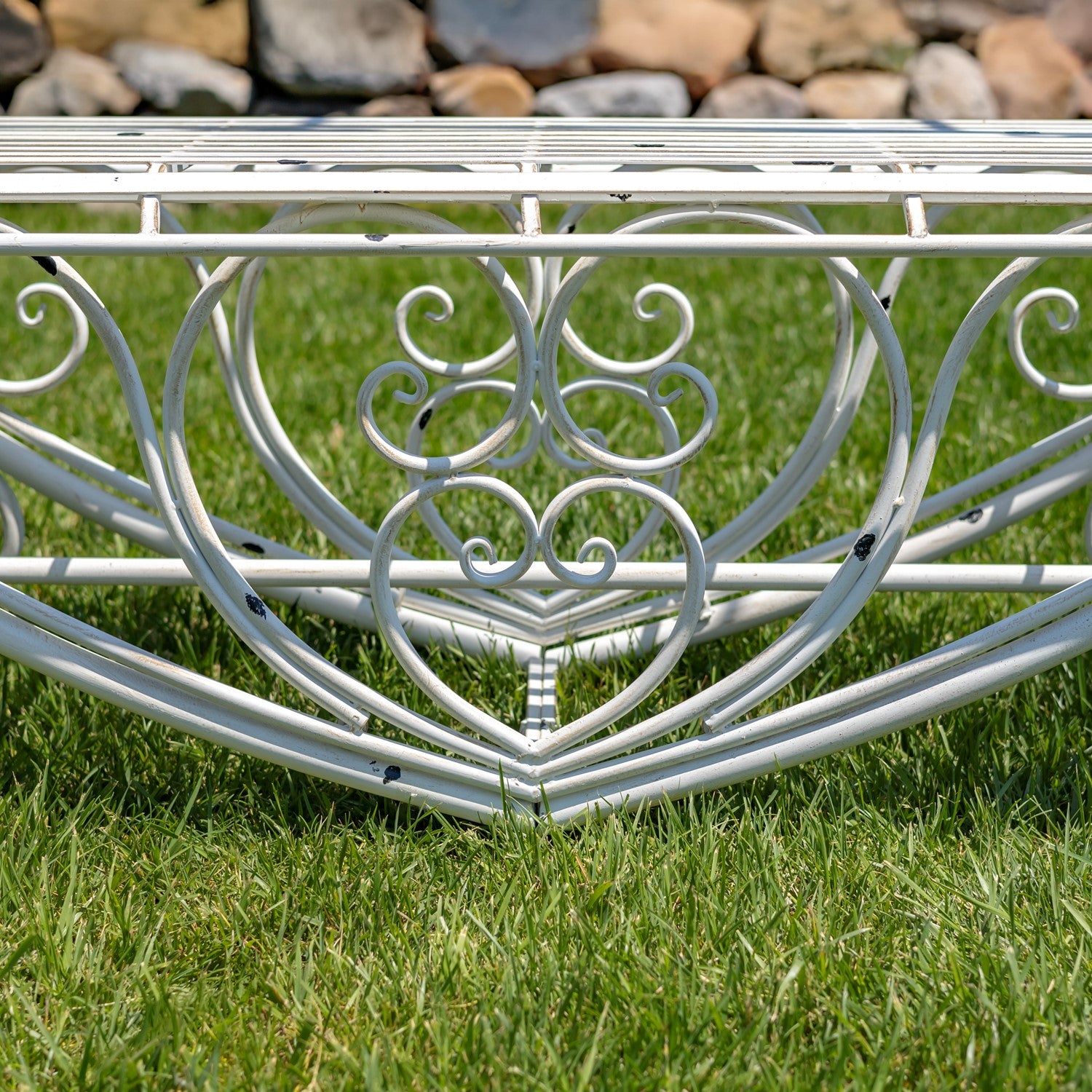 A white metal garden bench with an ornate scrollwork design and heart-shaped center motif, shown outdoors on green grass with rocks in the background.