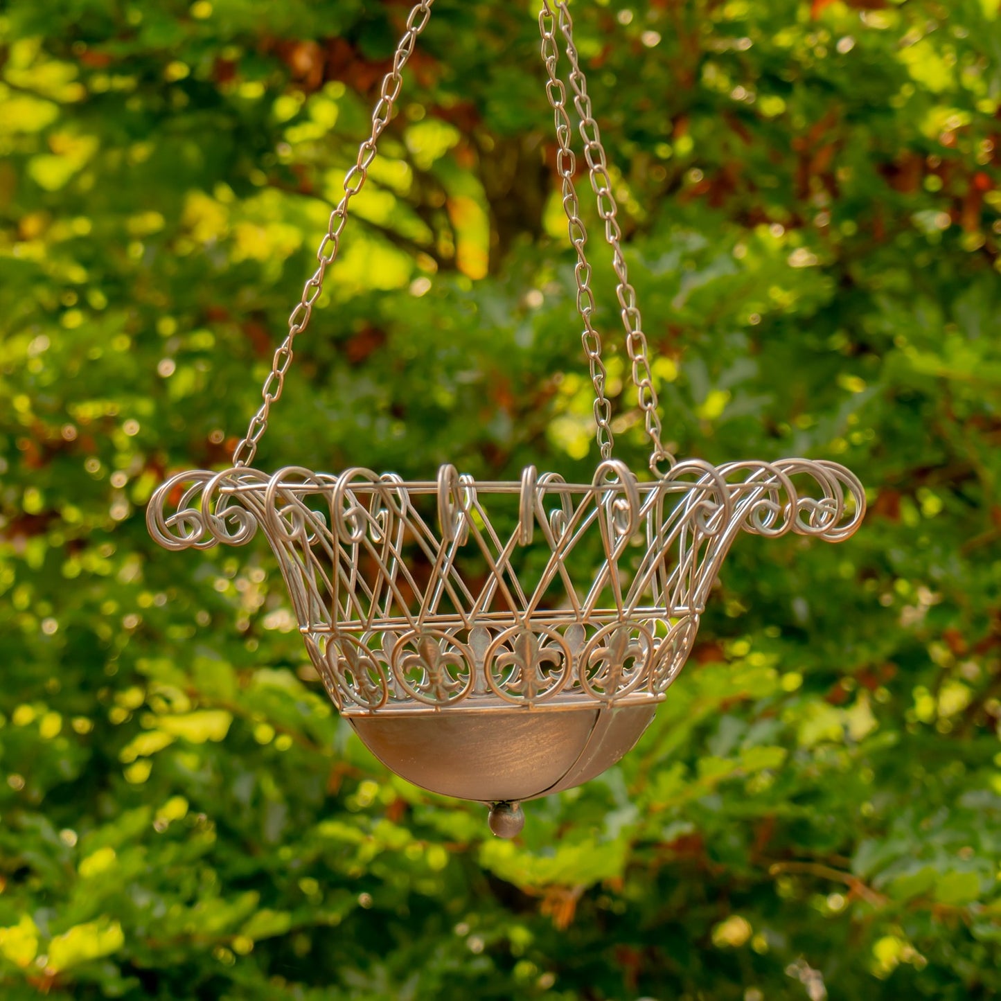 A hanging metal planter basket with an ornate scrollwork top and a shallow bowl below, suspended by chains against a blurred green garden background.