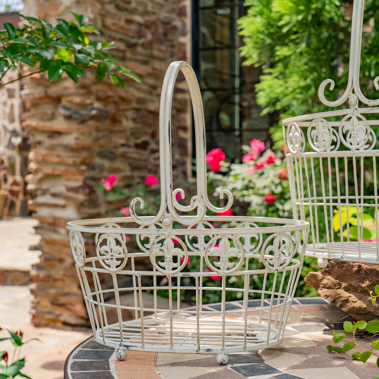 A white ornate metal garden basket with a tall handle sits on a stone patio table outdoors, with flowers and greenery blurred in the background.