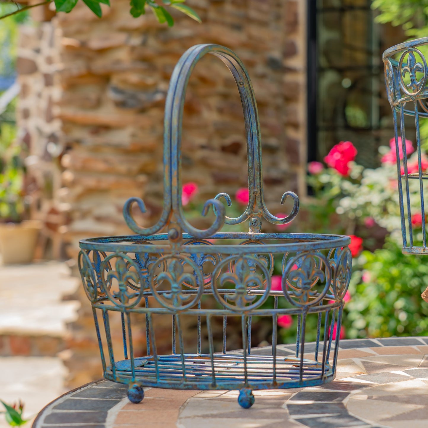 A decorative metal basket with a distressed blue and green patina finish, featuring an arched handle and circular cutout patterns, set on a stone patio with a blurred garden background.