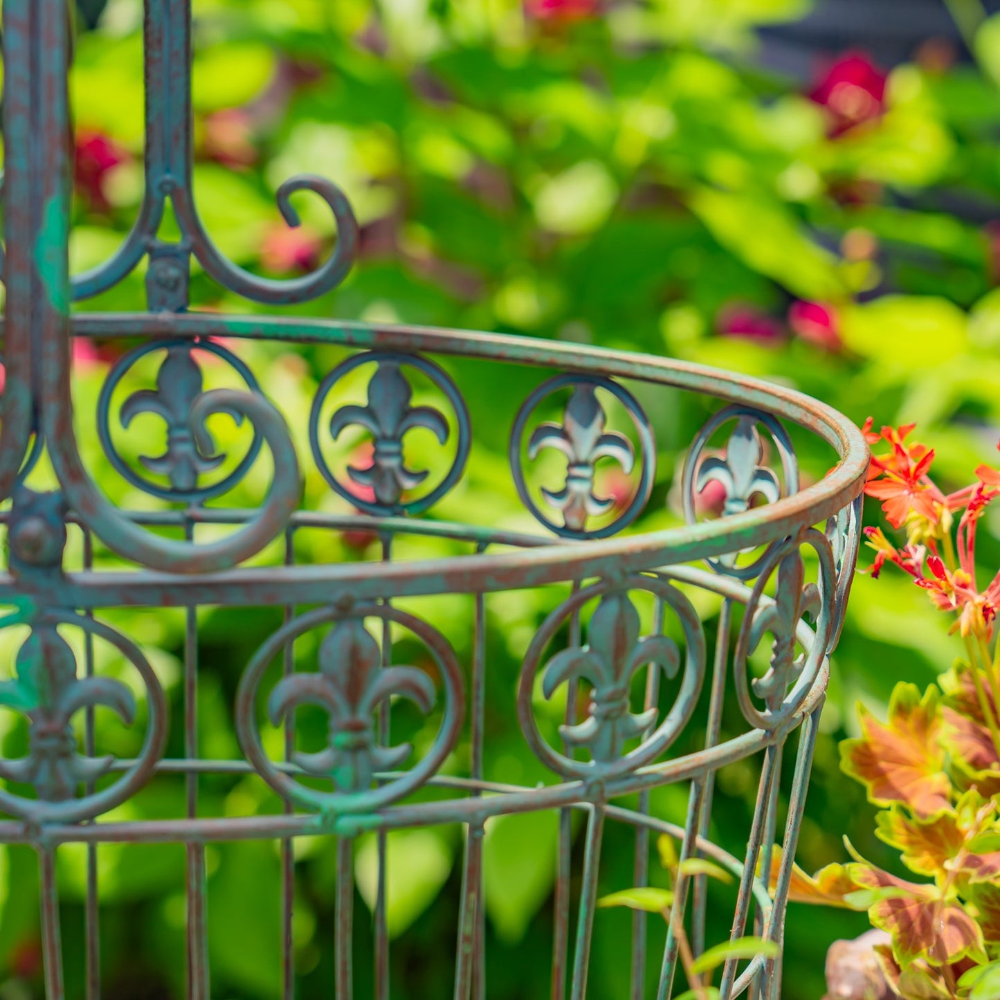 A close-up of an ornate metal garden stake or trellis section with a distressed teal-green patina and decorative scrollwork, set against a blurred garden background with green foliage and small red flowers.