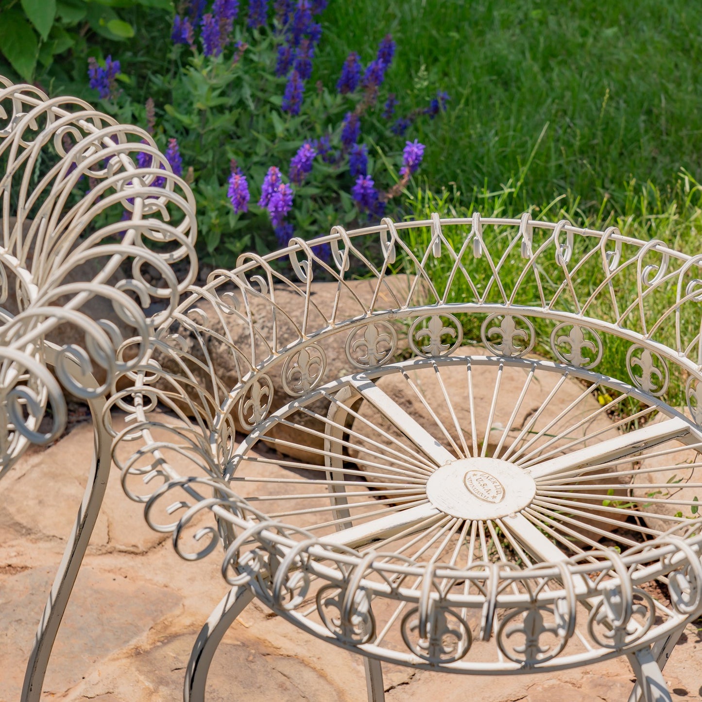 A close-up of a white metal garden chair with an ornate wheel-spoke design, set on a stone patio with green grass and purple flowers in the background.