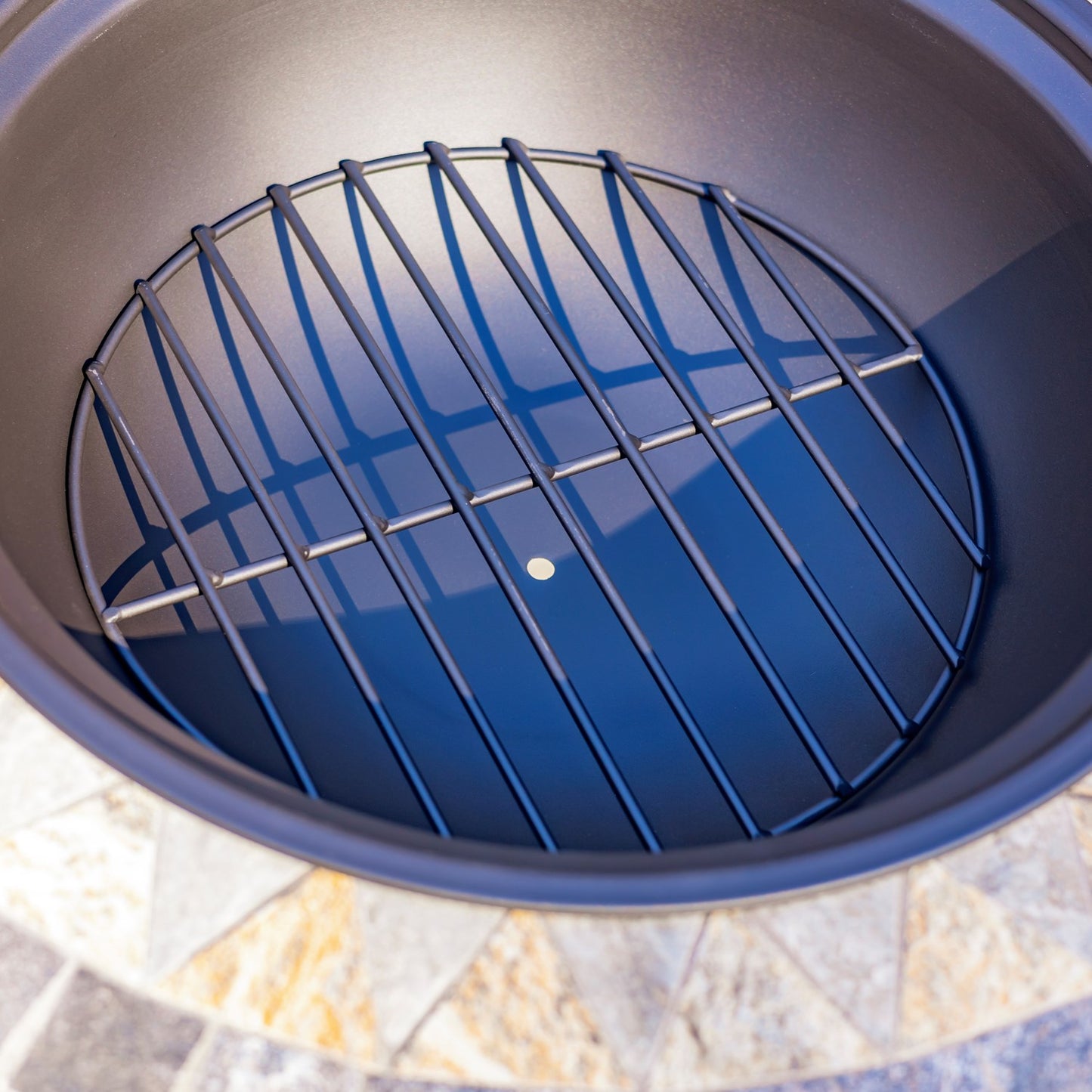 A round black metal grill grate insert resting inside a dark round grill pot, shown from a top-down view with a stone tile surface visible around the rim.