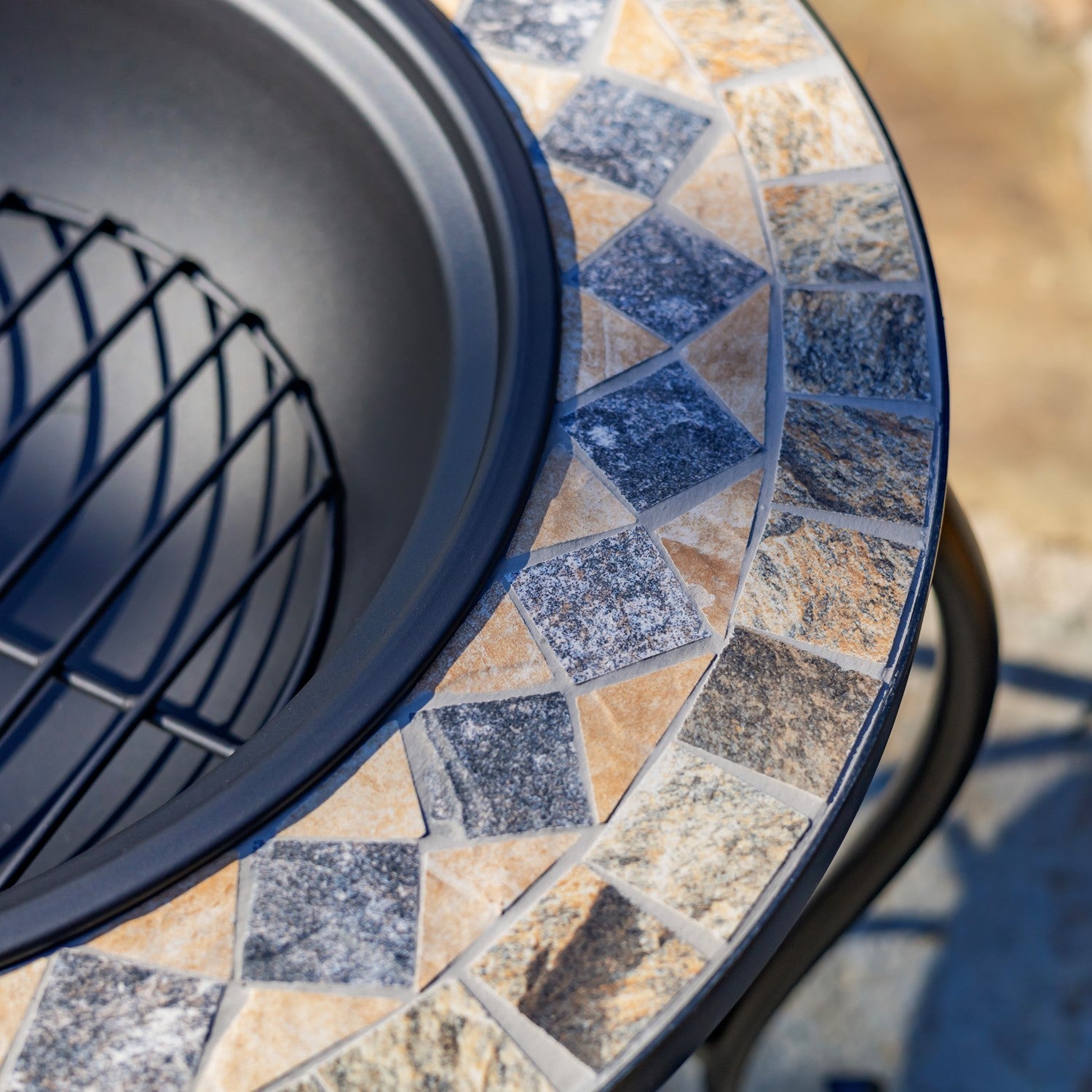 Close-up of a round outdoor fire pit table with a dark gray metal bowl and a mosaic tile rim featuring blue, beige, and brown stone pieces.