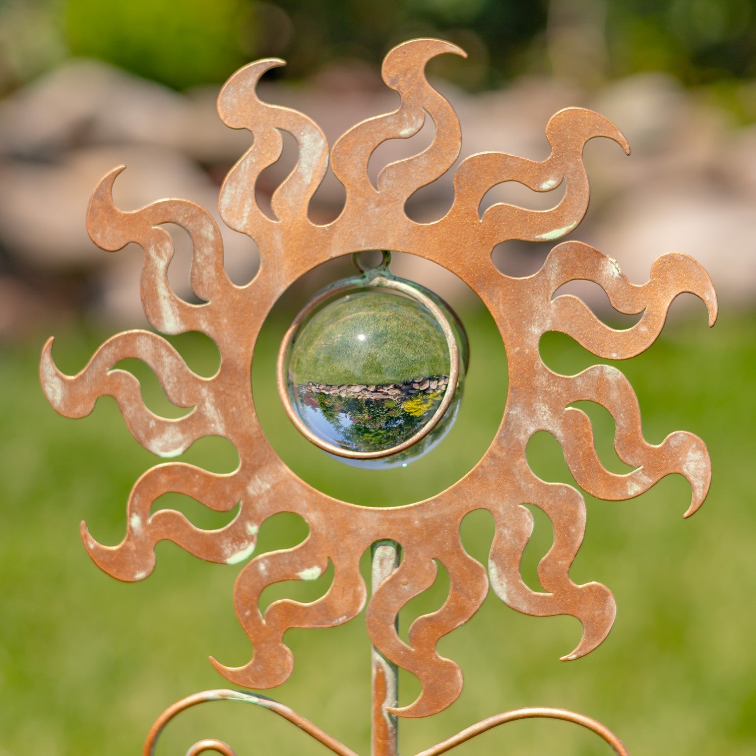 A copper-colored metal sun garden stake with flame-like rays and a clear glass sphere in the center, photographed outdoors against a blurred green garden background.