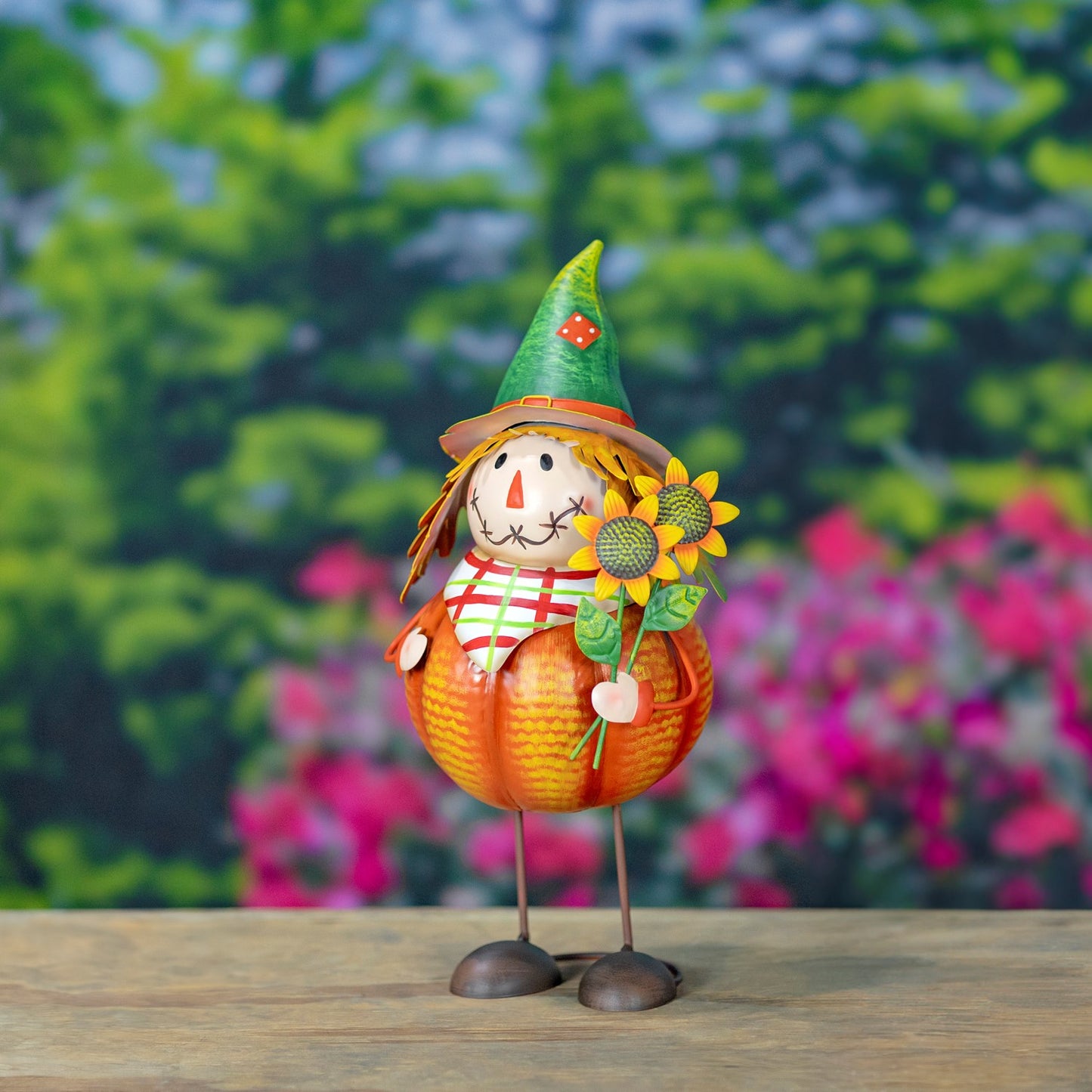 A decorative autumn scarecrow figurine standing on a wooden surface with a blurred garden background. The scarecrow has a pumpkin-shaped body, a green pointed hat, a striped shirt, and holds a sunflower.
