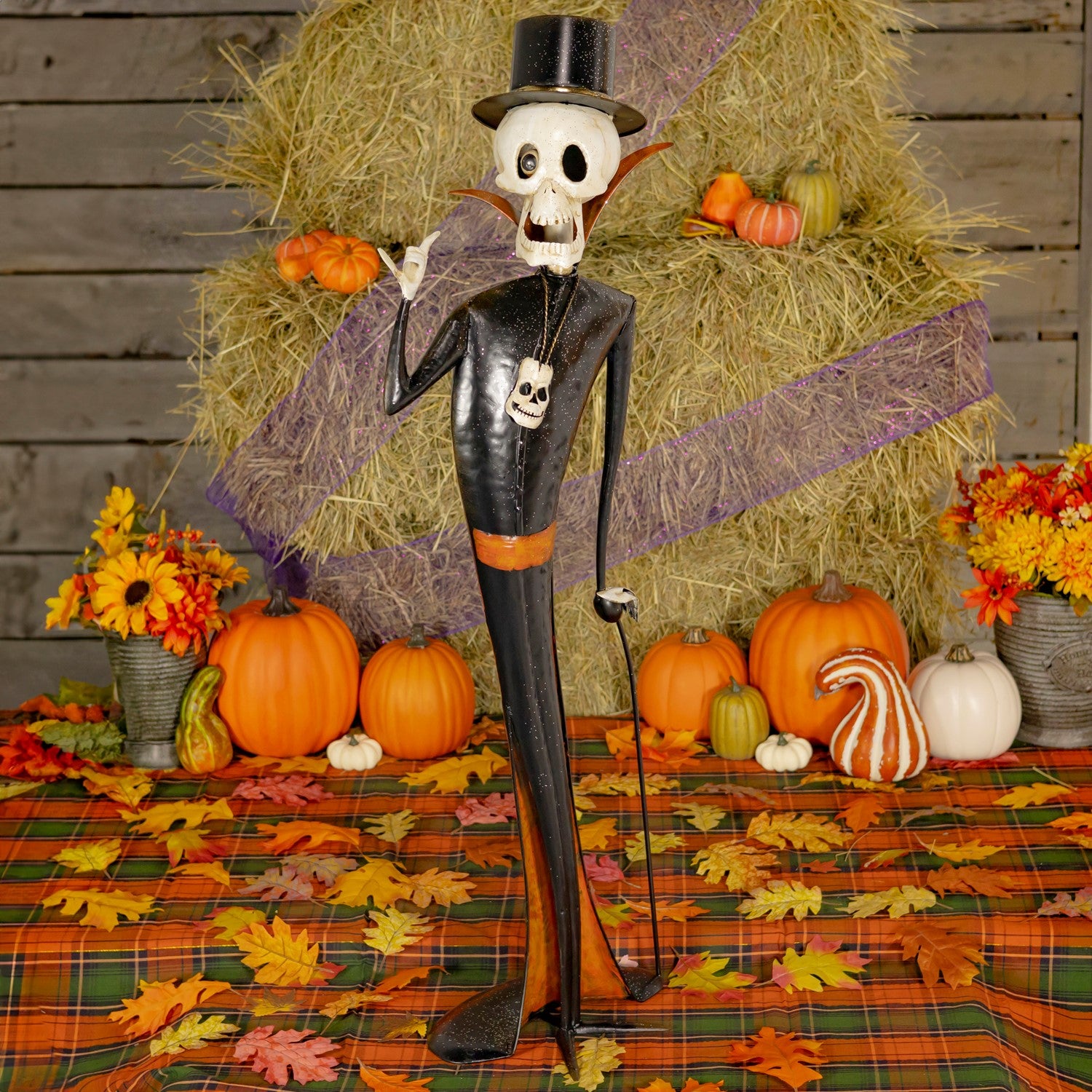 One male Halloween skeleton figurine dressed in black outfits, standing on a plaid autumn tablecloth with pumpkins, flowers, and a hay bale backdrop.