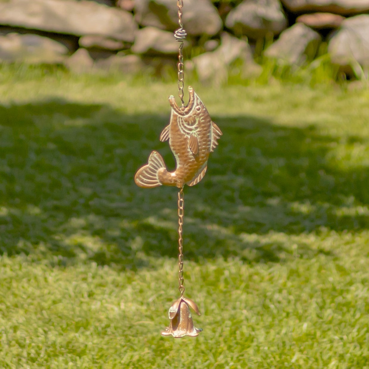 Decorative fish-shaped wind chime hanging on grass with a stone wall in the background