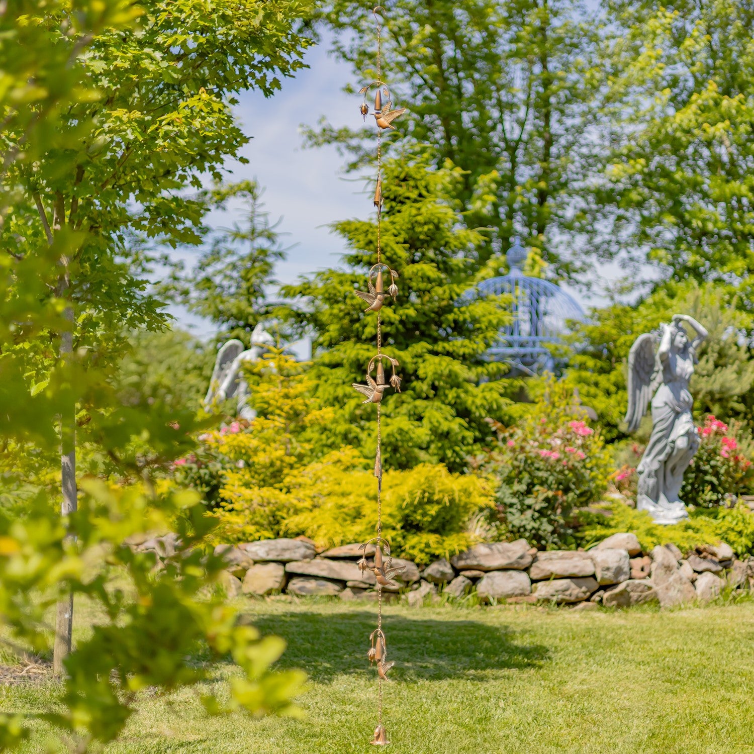 A hanging metal garden wind spinner with birdcage details, suspended in a lush green backyard garden with stone walls, angel statues, and a blue gazebo in the background.