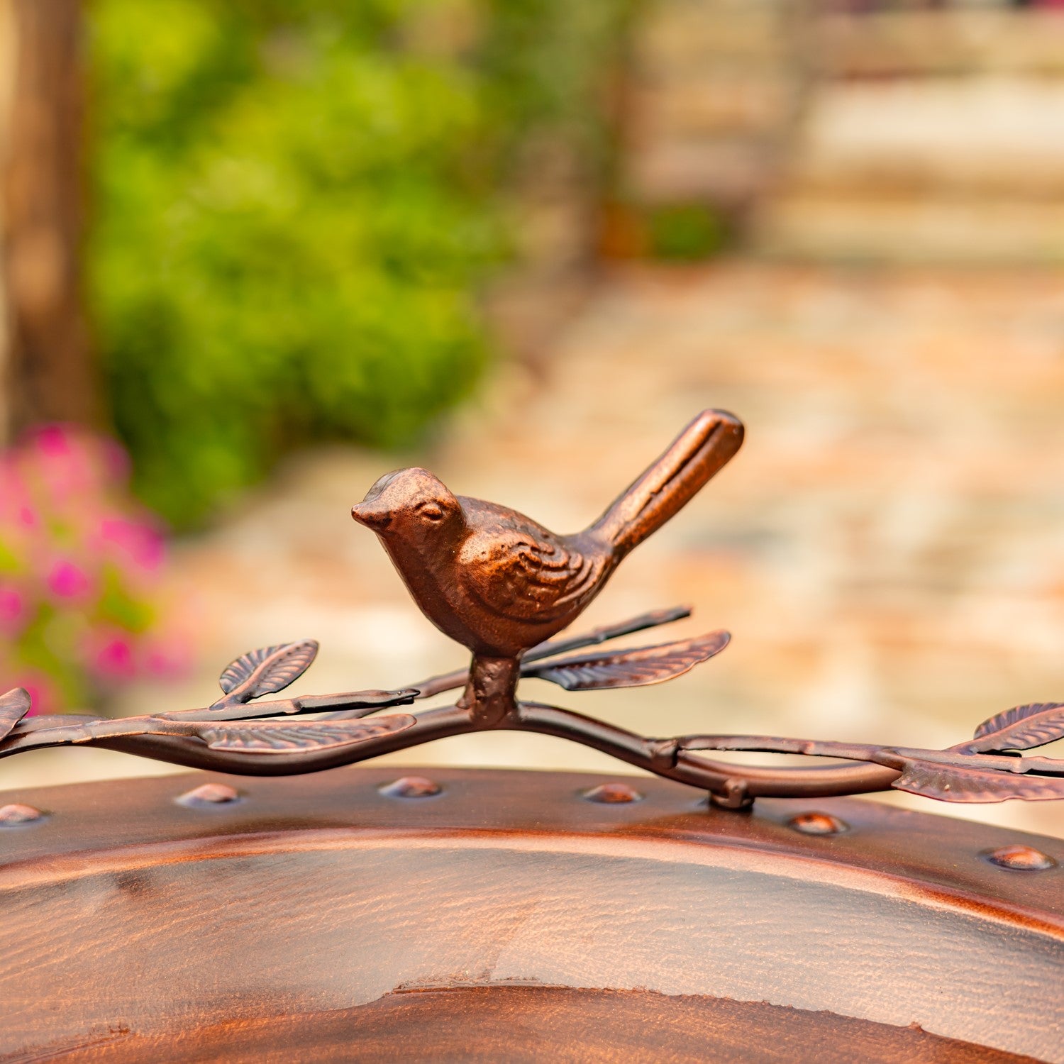 A small bird figurine in a bronze finish perched on a leafy metal branch, mounted on the edge of a round wooden garden table with a blurred outdoor garden background.