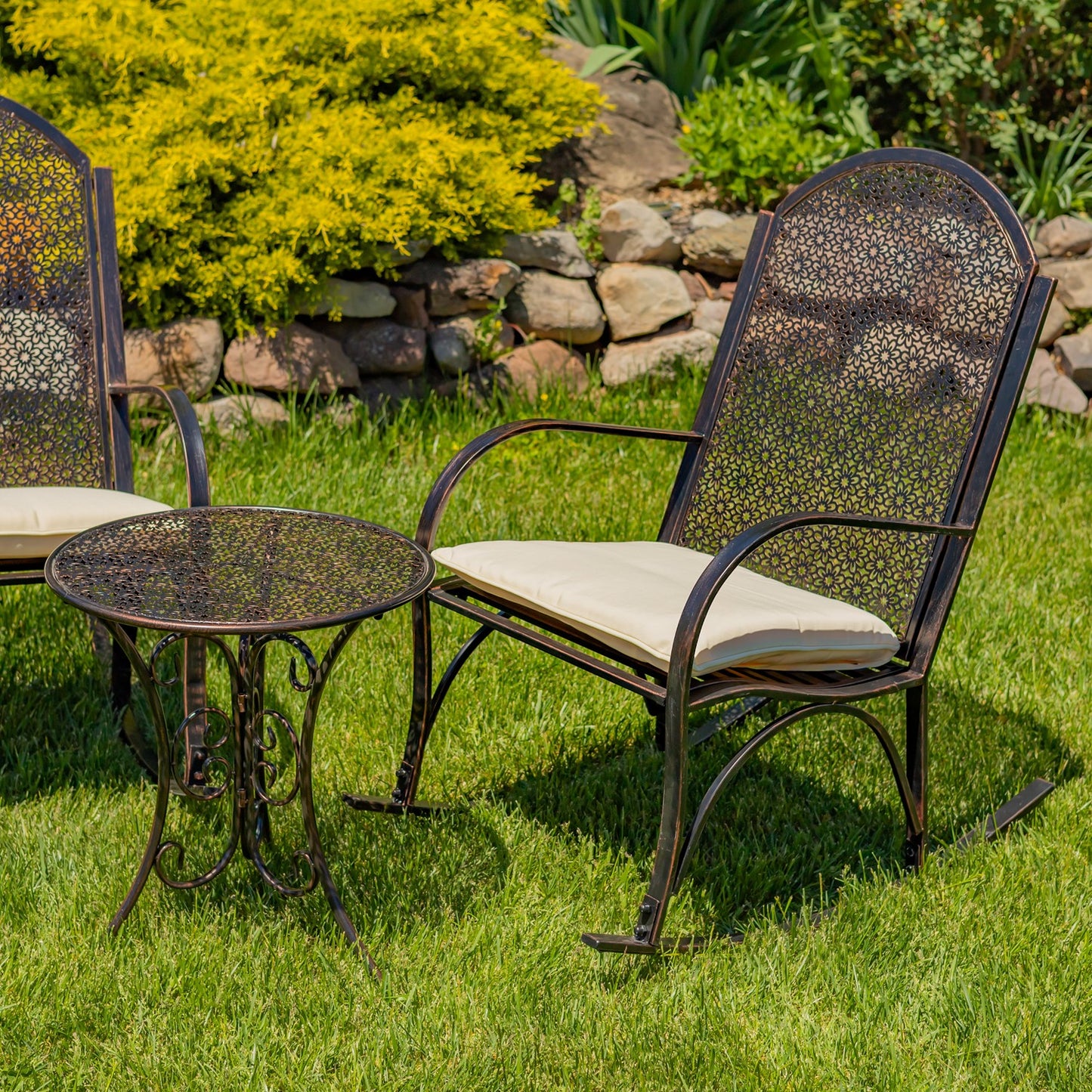 Outdoor patio furniture set on a green lawn, featuring a dark metal armchair with a cream cushion, a matching side table, and part of another chair in the background near yellow-green shrubs and a stone wall.
