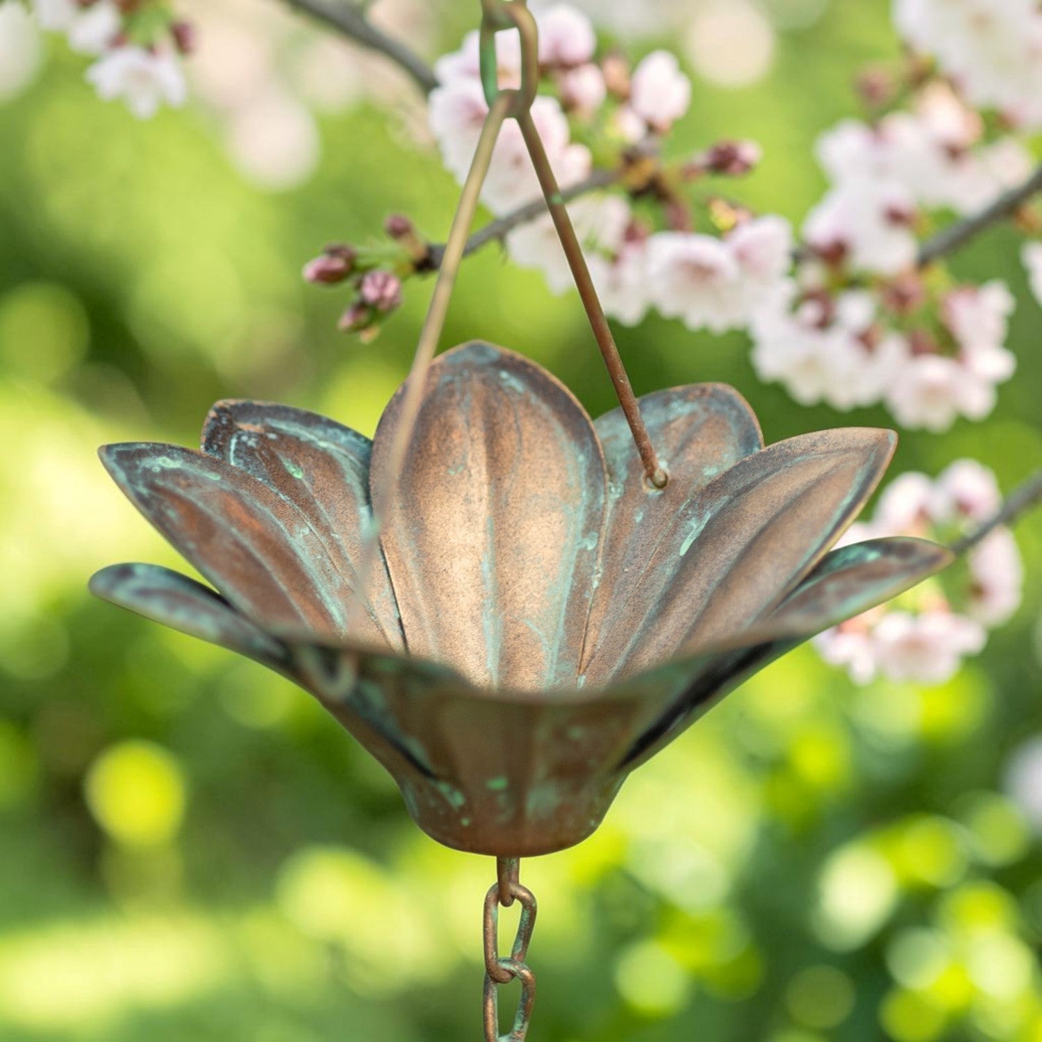 A bronze metal flower-shaped garden wind spinner hanging from a thin rod, set against a soft-focus background of green foliage and light pink blossoms.