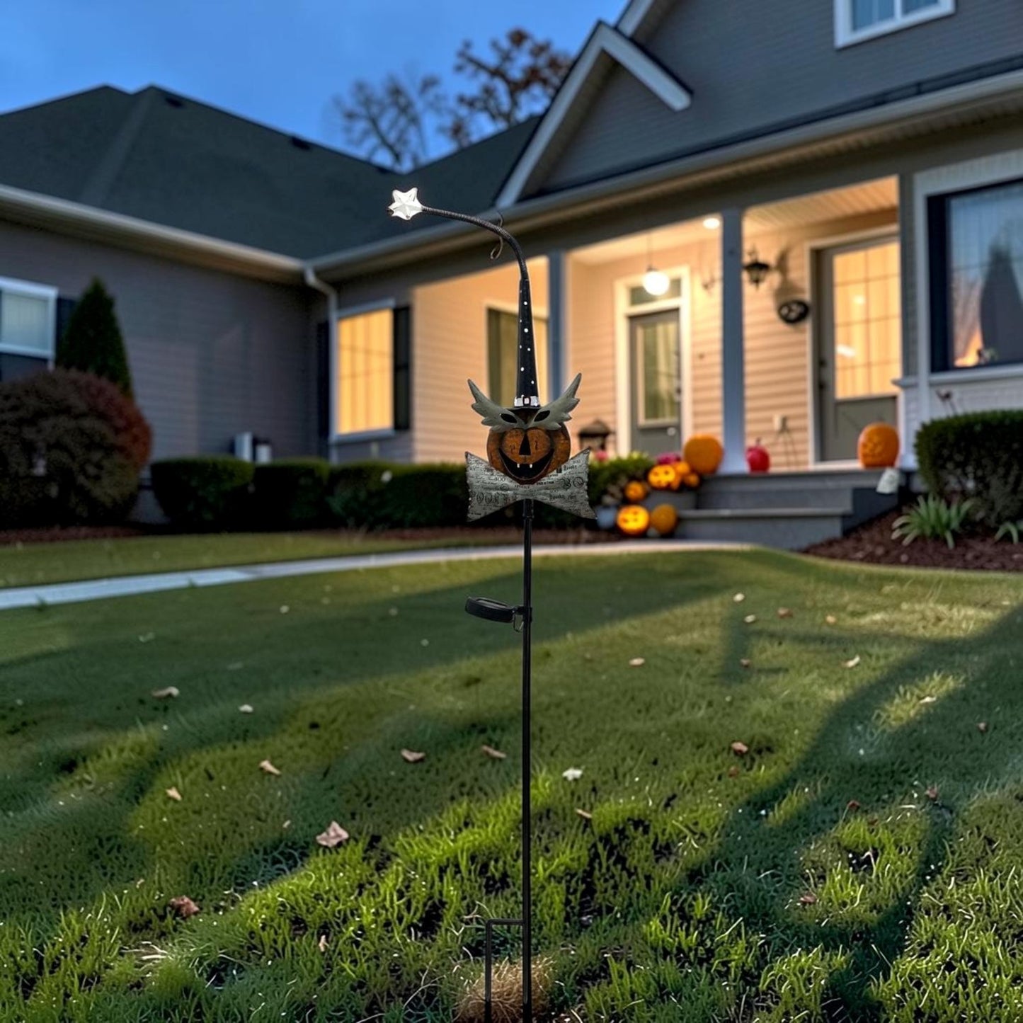 A Halloween yard stake garden light featuring a jack-o'-lantern face, bat wings, and a star-topped metal wire arm, shown lit in a front yard with a house and pumpkins in the background.