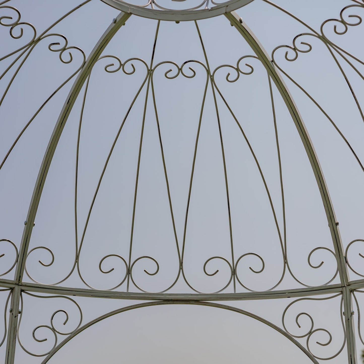 Close-up of an ornate metal gazebo or pergola frame with curved scrollwork and vertical support beams against a pale sky background.