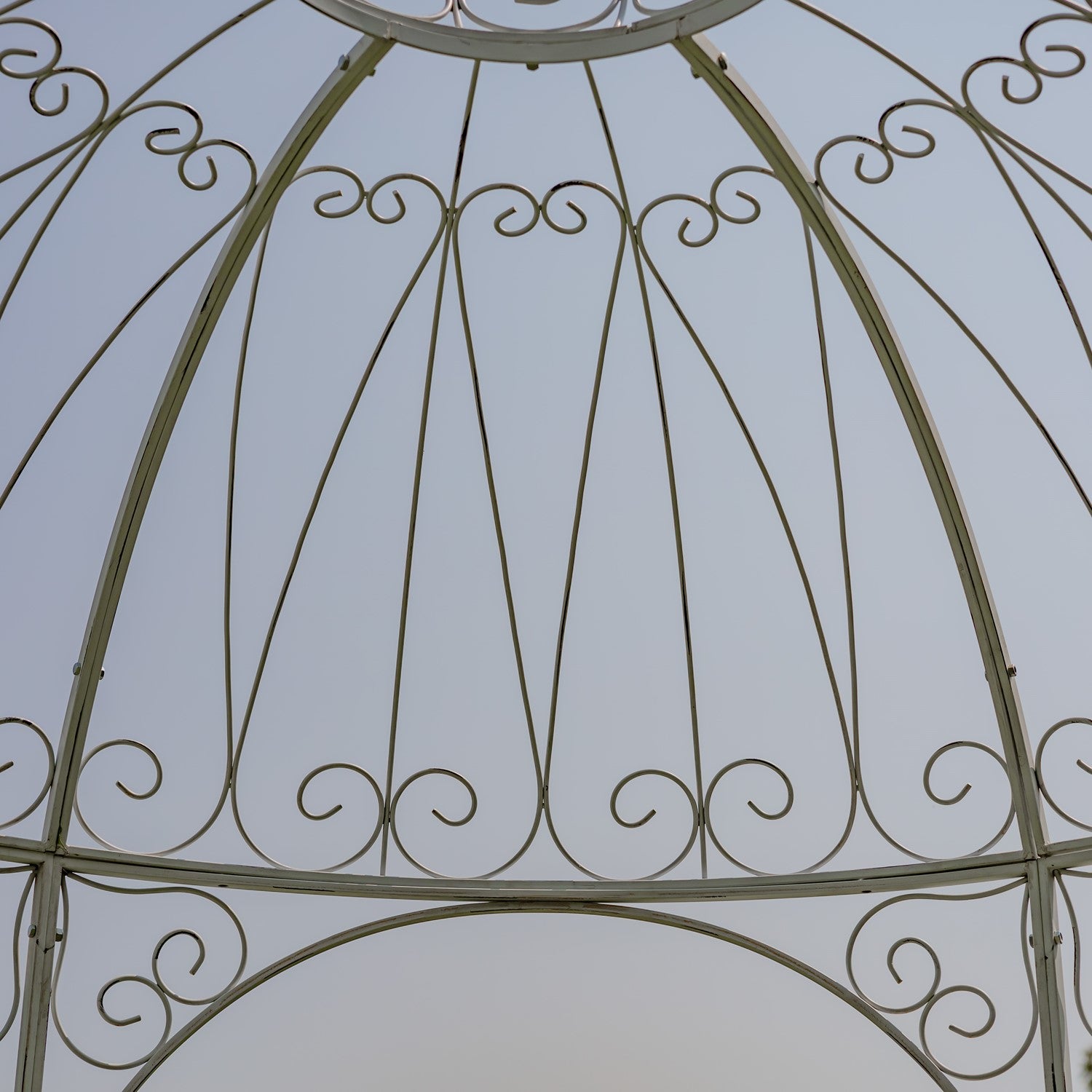 Close-up of an ornate metal gazebo or pergola frame with curved scrollwork and vertical support beams against a pale sky background.