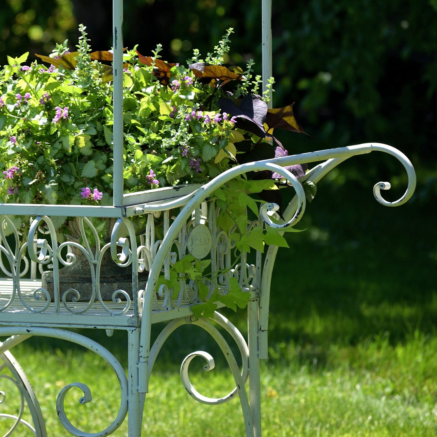 A white wrought iron garden planter cart filled with green foliage and small purple flowers, set outdoors on a grassy lawn with a blurred green background.