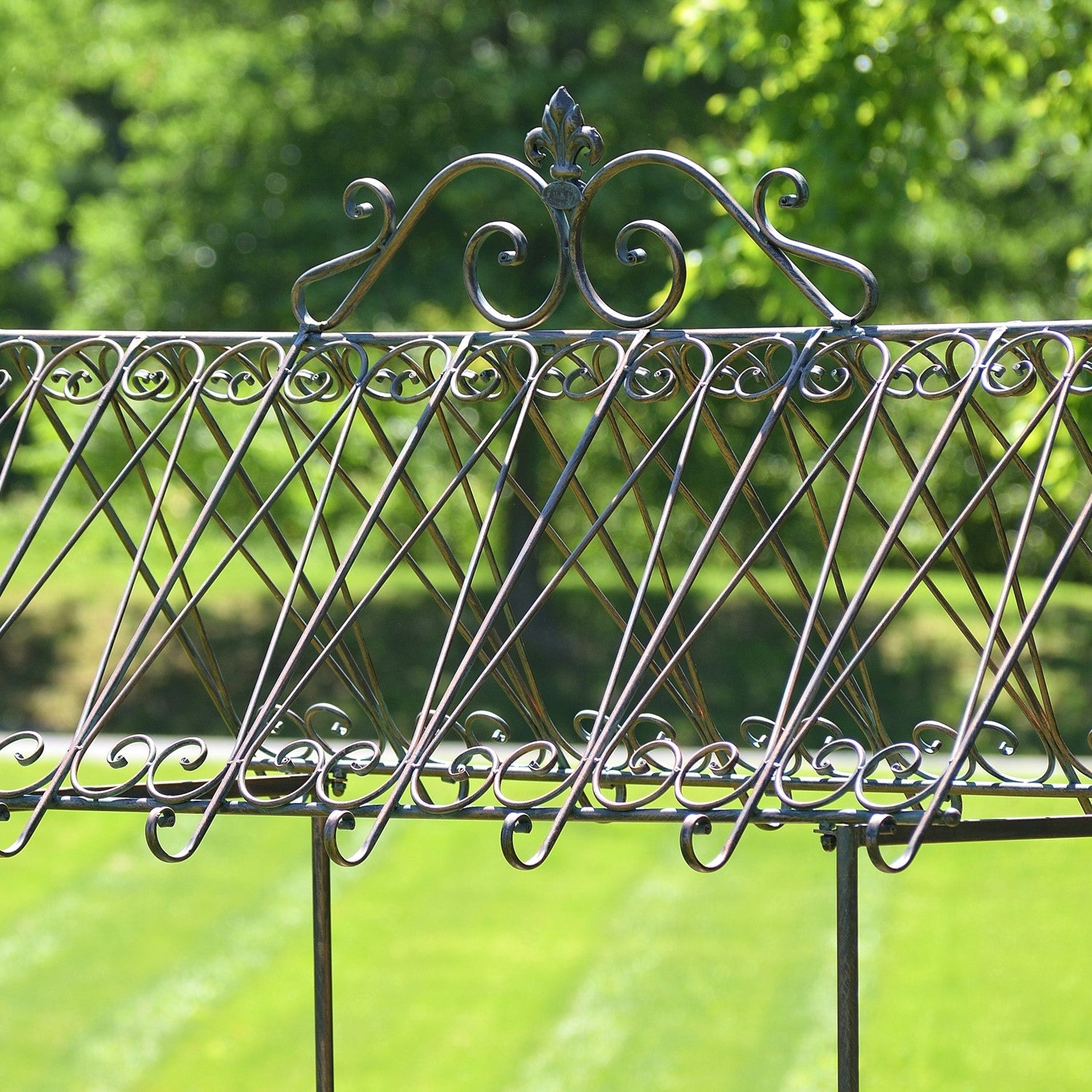 A close-up of an ornate wrought iron fence or gate with decorative scrollwork and a diamond lattice pattern, set against a blurred green garden background.