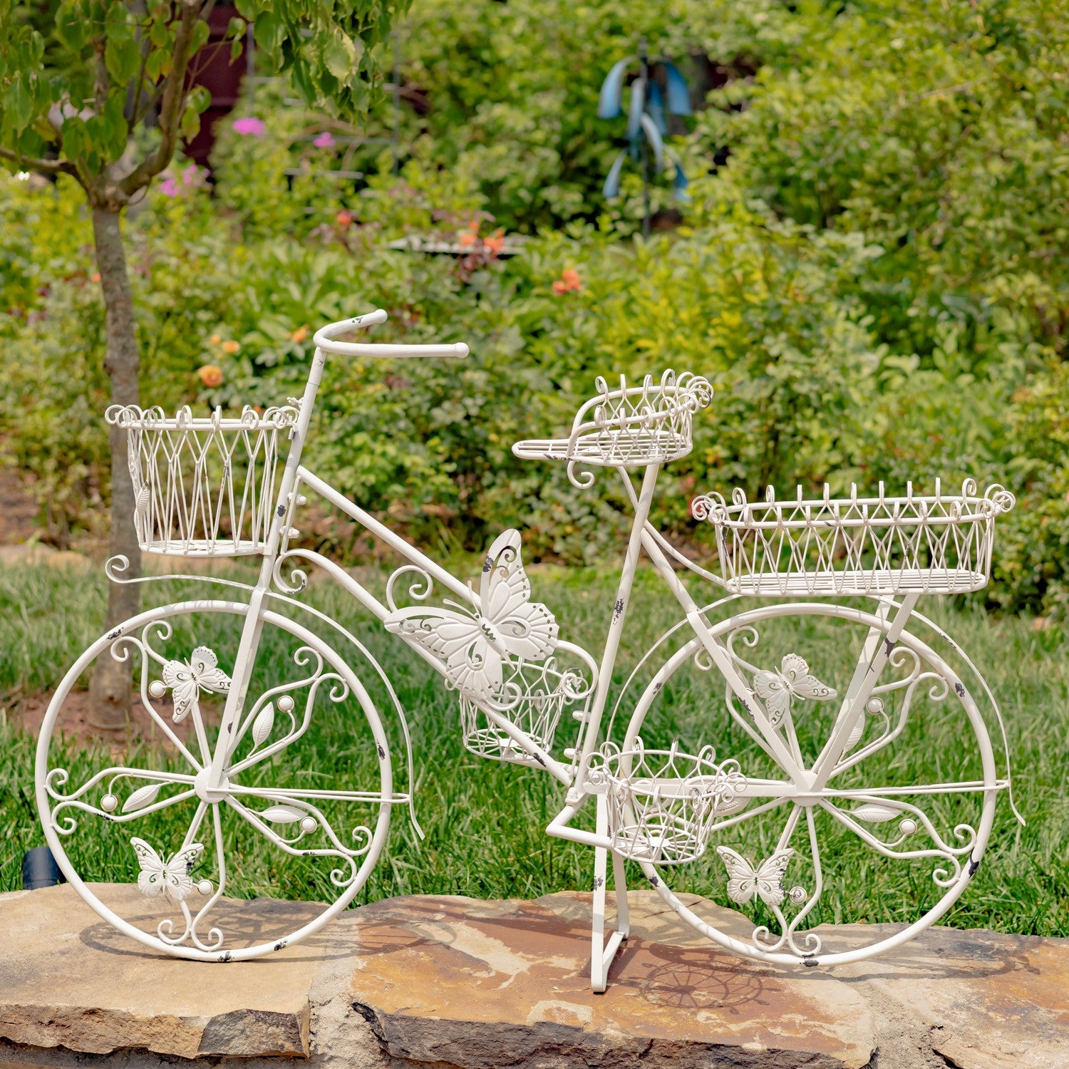 A decorative white metal garden bicycle sculpture with butterfly and floral details, displayed outdoors on a stone slab with green foliage in the background.